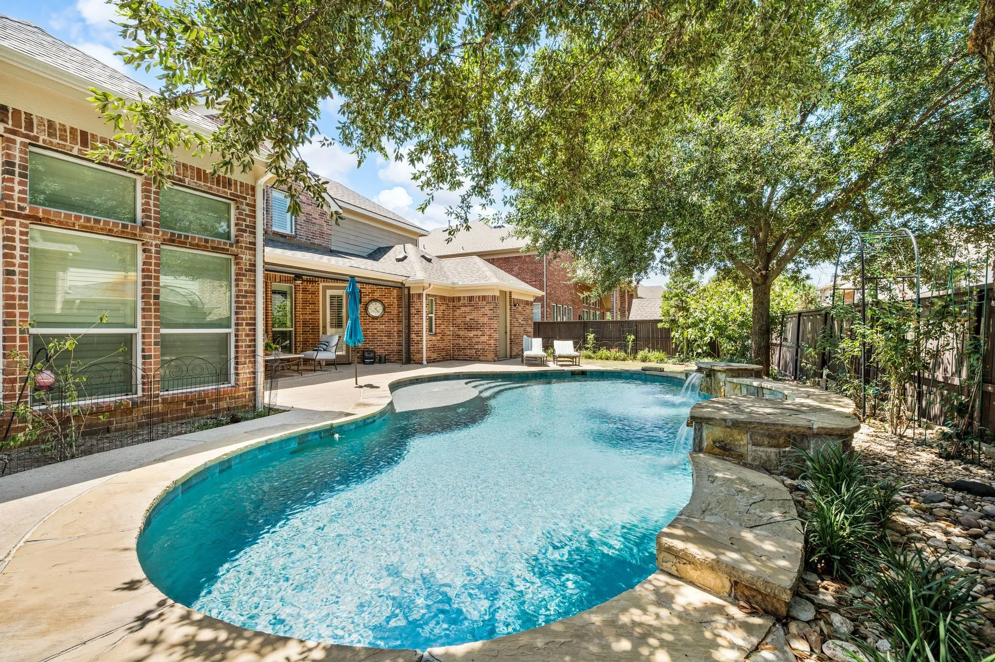 Freeform swimming pool with a stone waterfall feature, expansive patio, and mature trees.