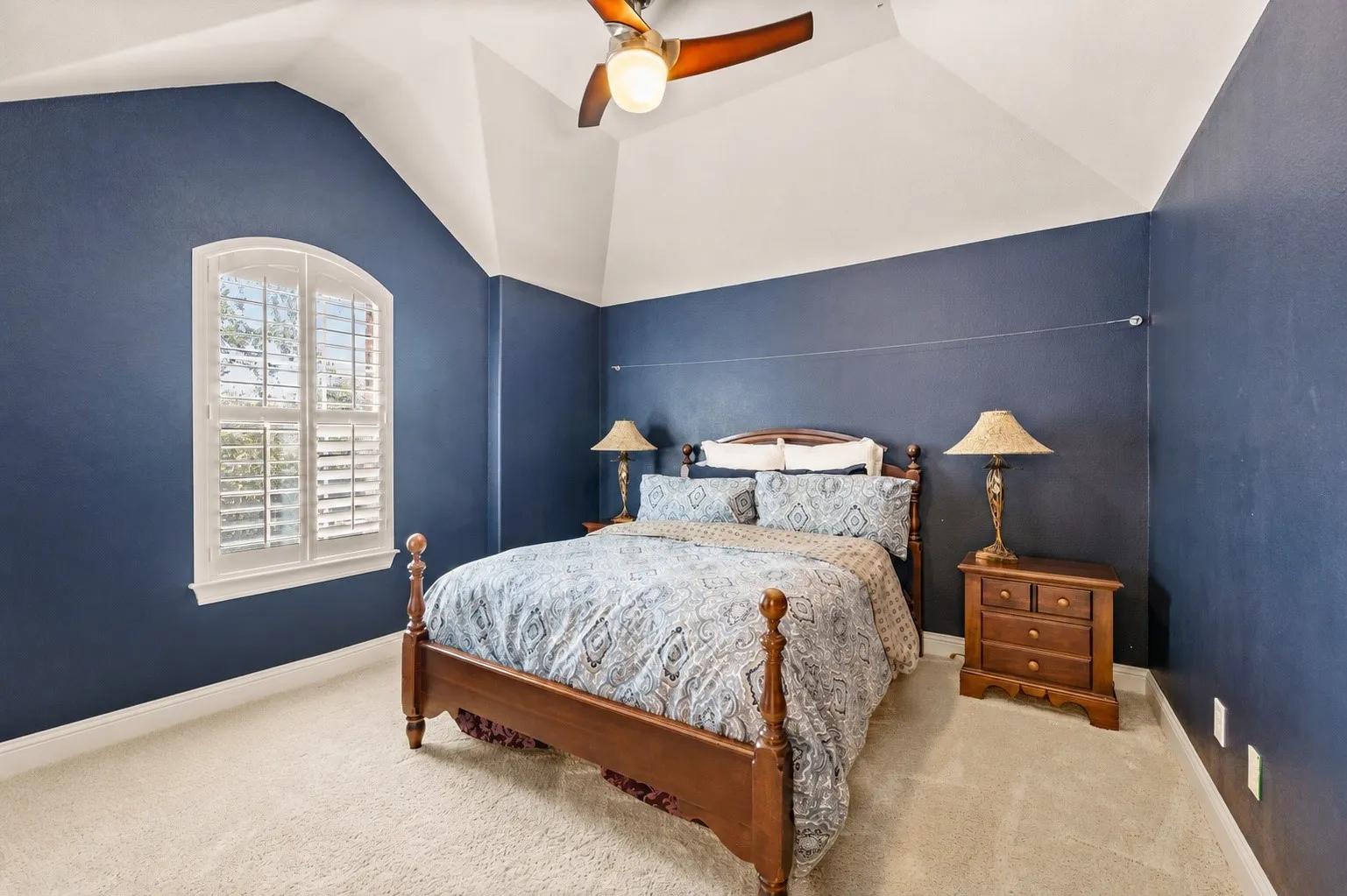 Arched window with plantation shutters, vaulted ceiling, ceiling fan with light fixture, and plush carpeting