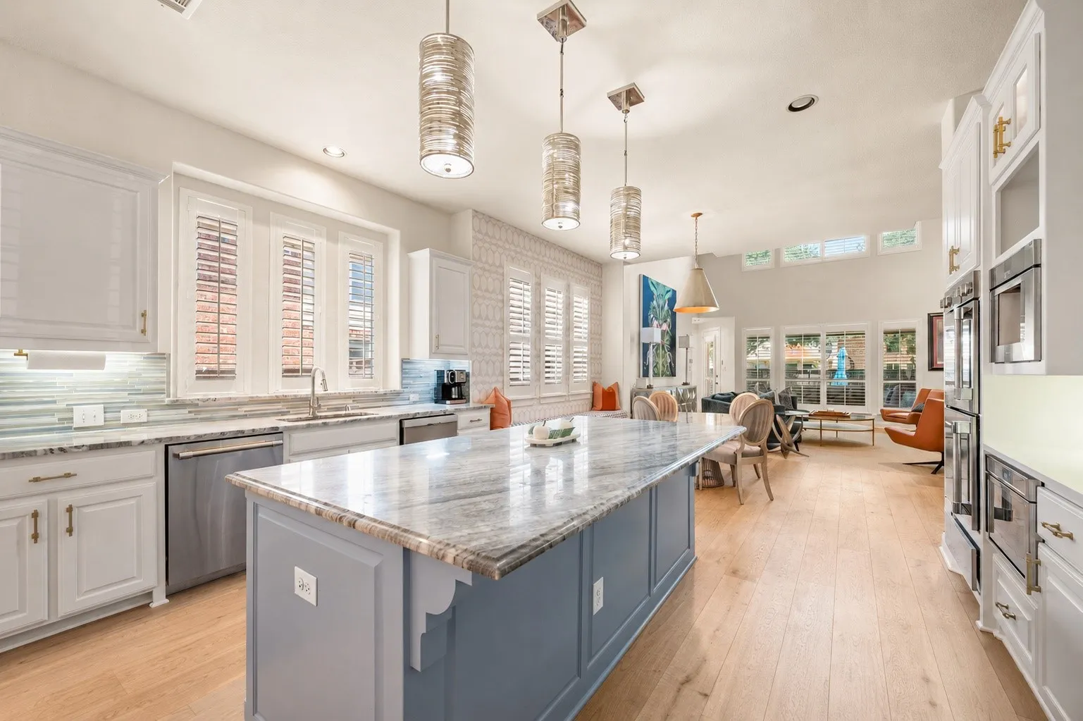 Spacious kitchen featuring an expansive center island with marble countertop, light wood-finish flooring, white cabinetry with gold-tone hardware, and integrated Thermador stainless steel appliances