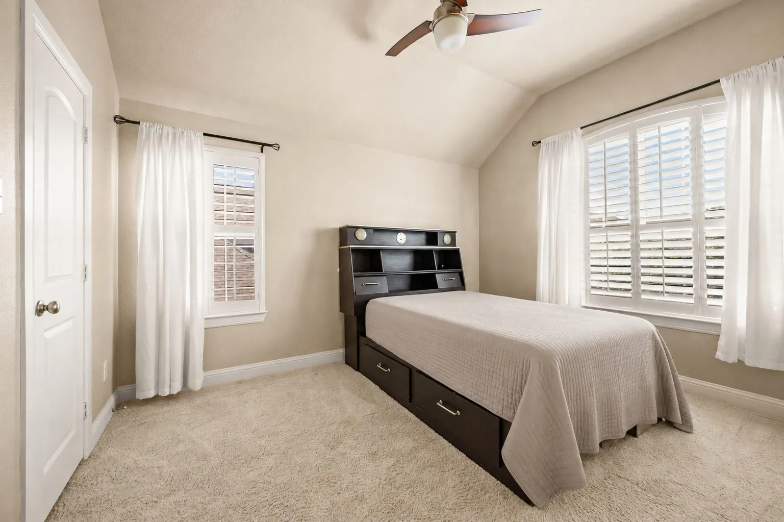 Vaulted ceiling with a ceiling fan, plush carpeting, two windows with plantation shutters, and a white paneled door