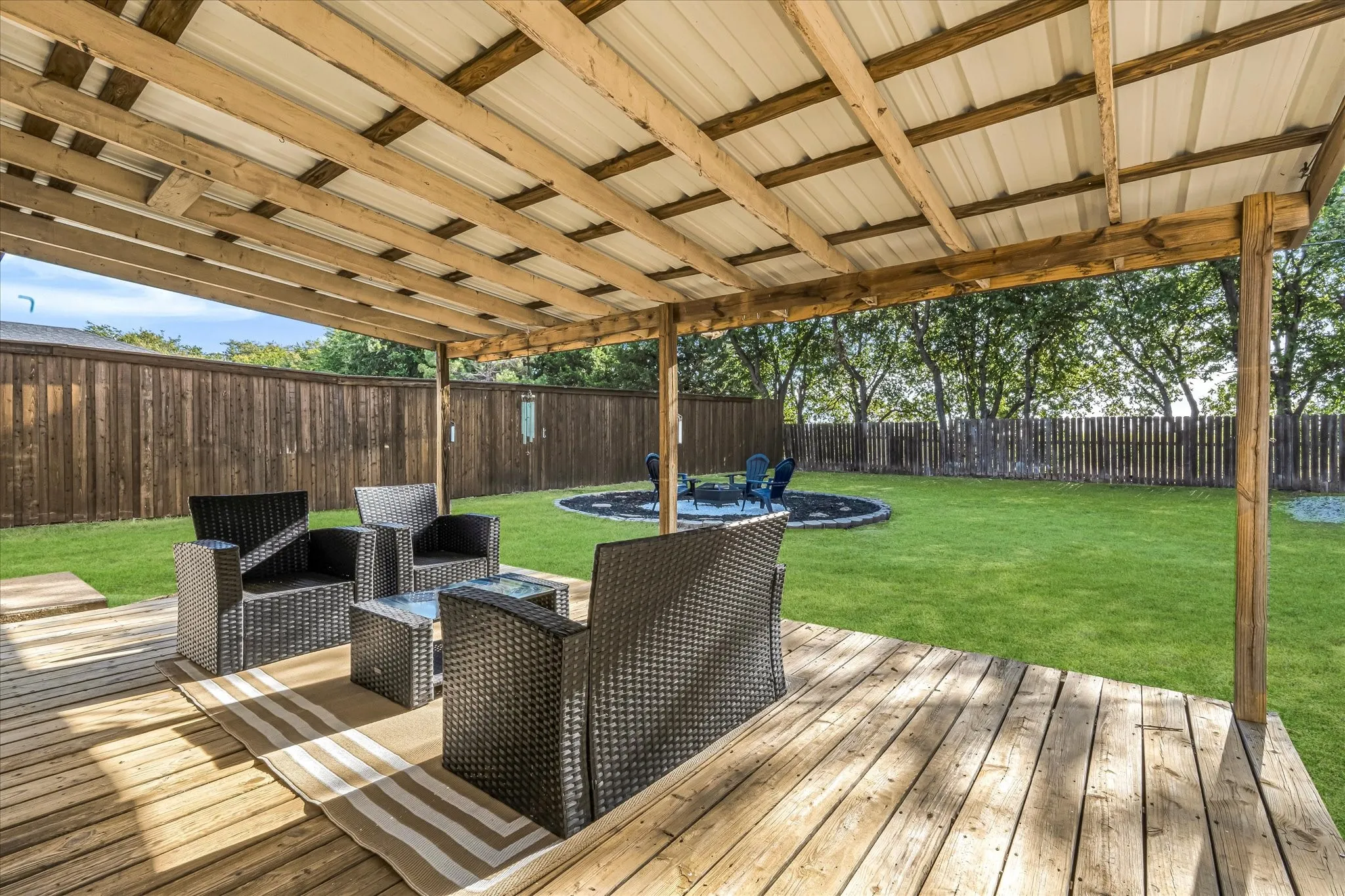 Expansive wood deck with a covered patio structure, lush green lawn, full wood privacy fence, and a circular fire pit area