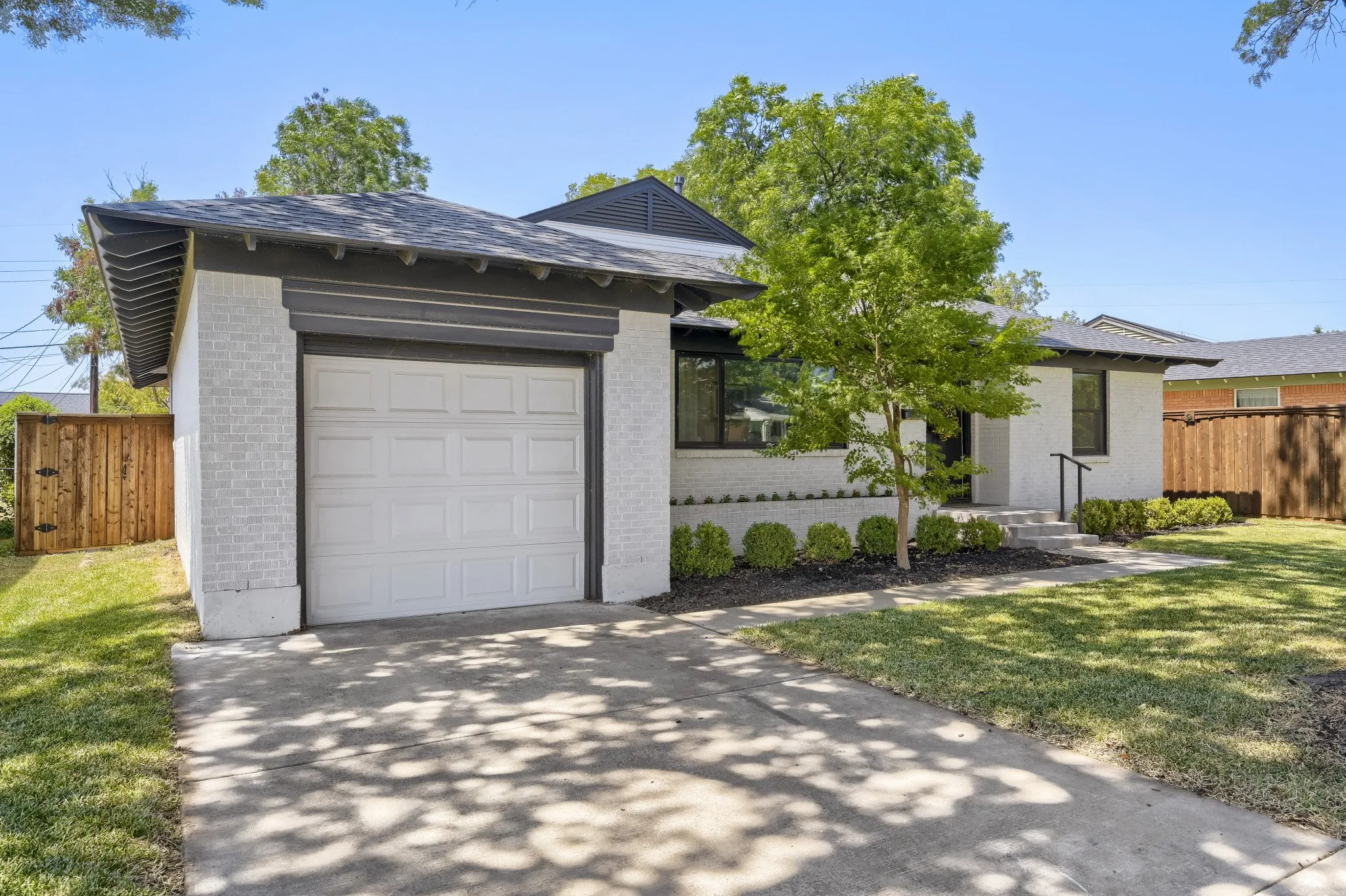 White brick exterior with dark trim accents, attached garage with a white panel door, mature landscaping, and a concrete driveway