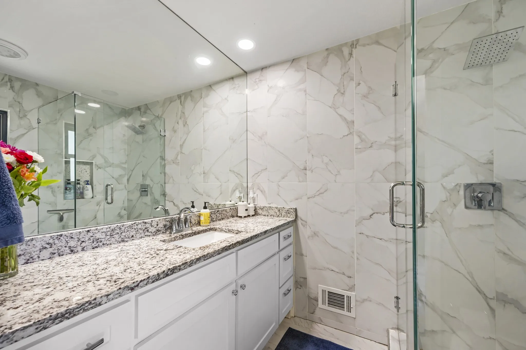 Expansive vanity with a light granite countertop and undermount sink, mirrored wall, and glass-enclosed shower with a rainfall showerhead