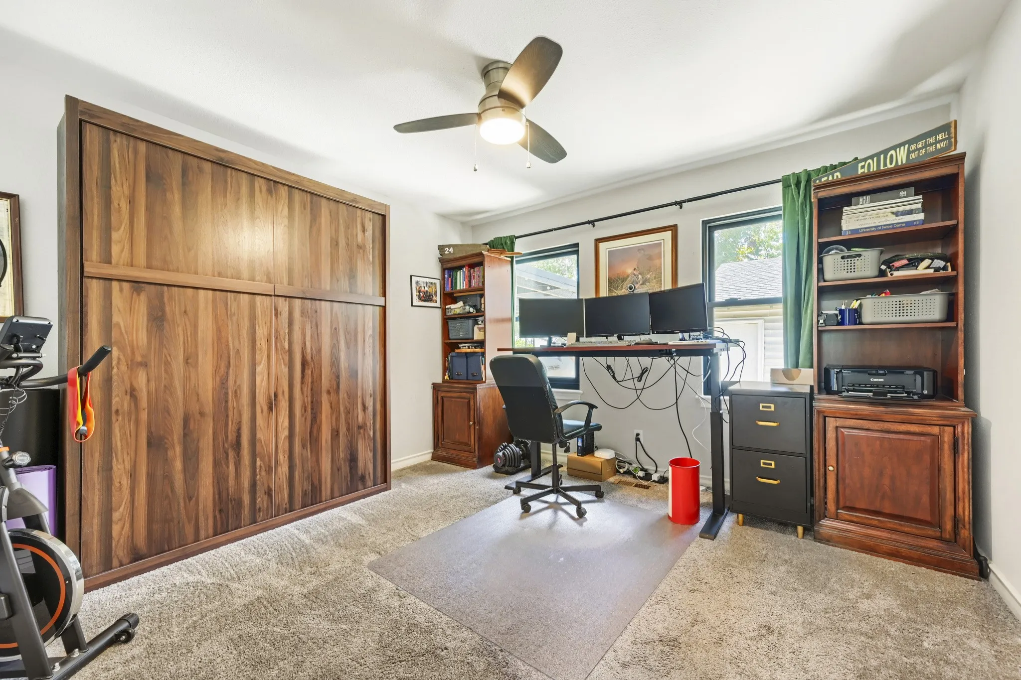 Built-in wood-finish wall unit, ceiling fan, multiple windows with dark trim, and a wood-finish bookshelf