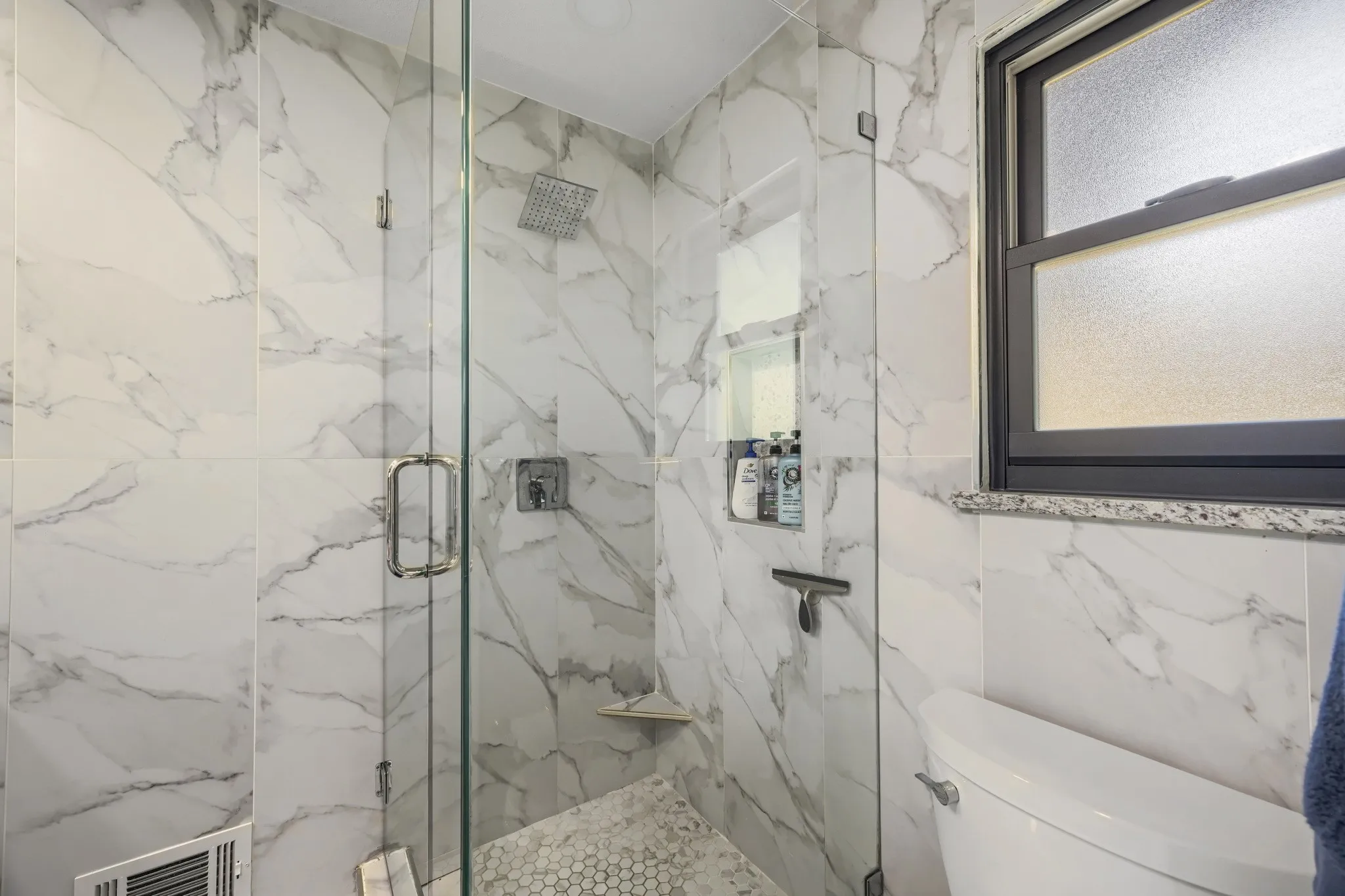 Walk-in shower with clear glass enclosure, a rain showerhead, white marble-look wall tiles, an integrated shower niche, and hexagonal floor tiles
