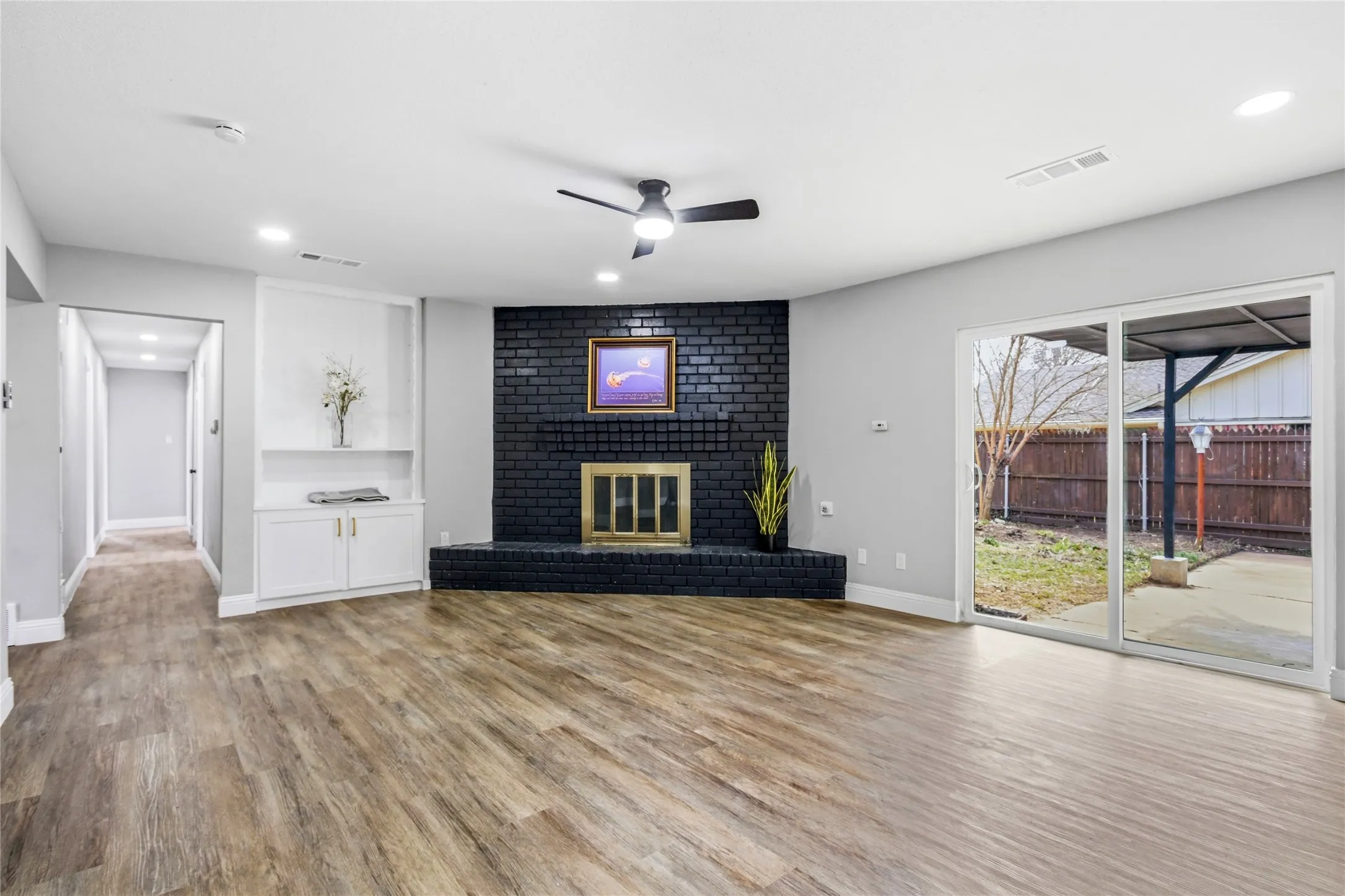 Spacious room featuring wood-finish flooring, a black brick fireplace with a brass firebox, built-in cabinetry, recessed lighting, and a sliding glass door