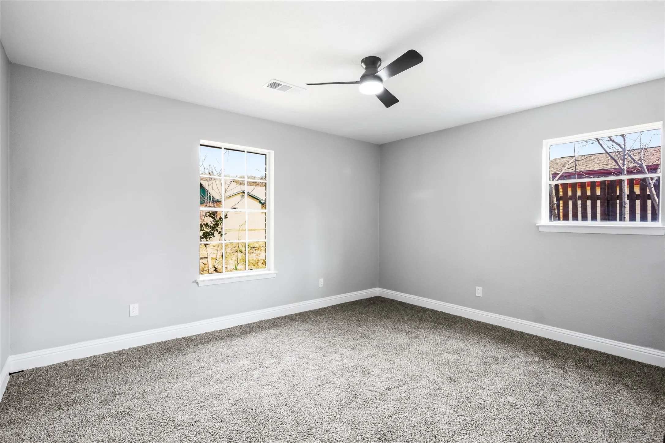Neutral-toned walls, two windows with white trim, a black ceiling fan with integrated lighting, and gray carpeted flooring