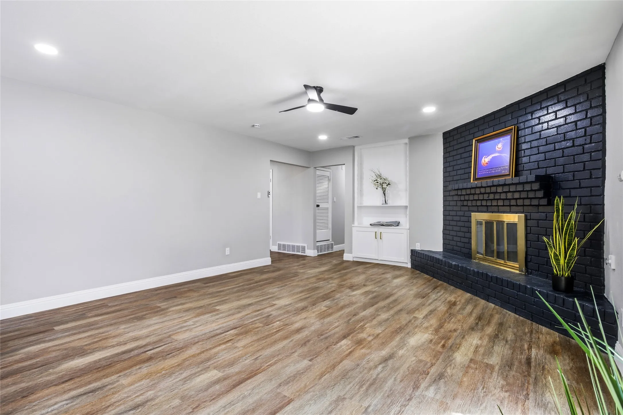 Wood-finish flooring, recessed lighting, ceiling fan, black brick fireplace with a gold-tone insert, and built-in cabinetry