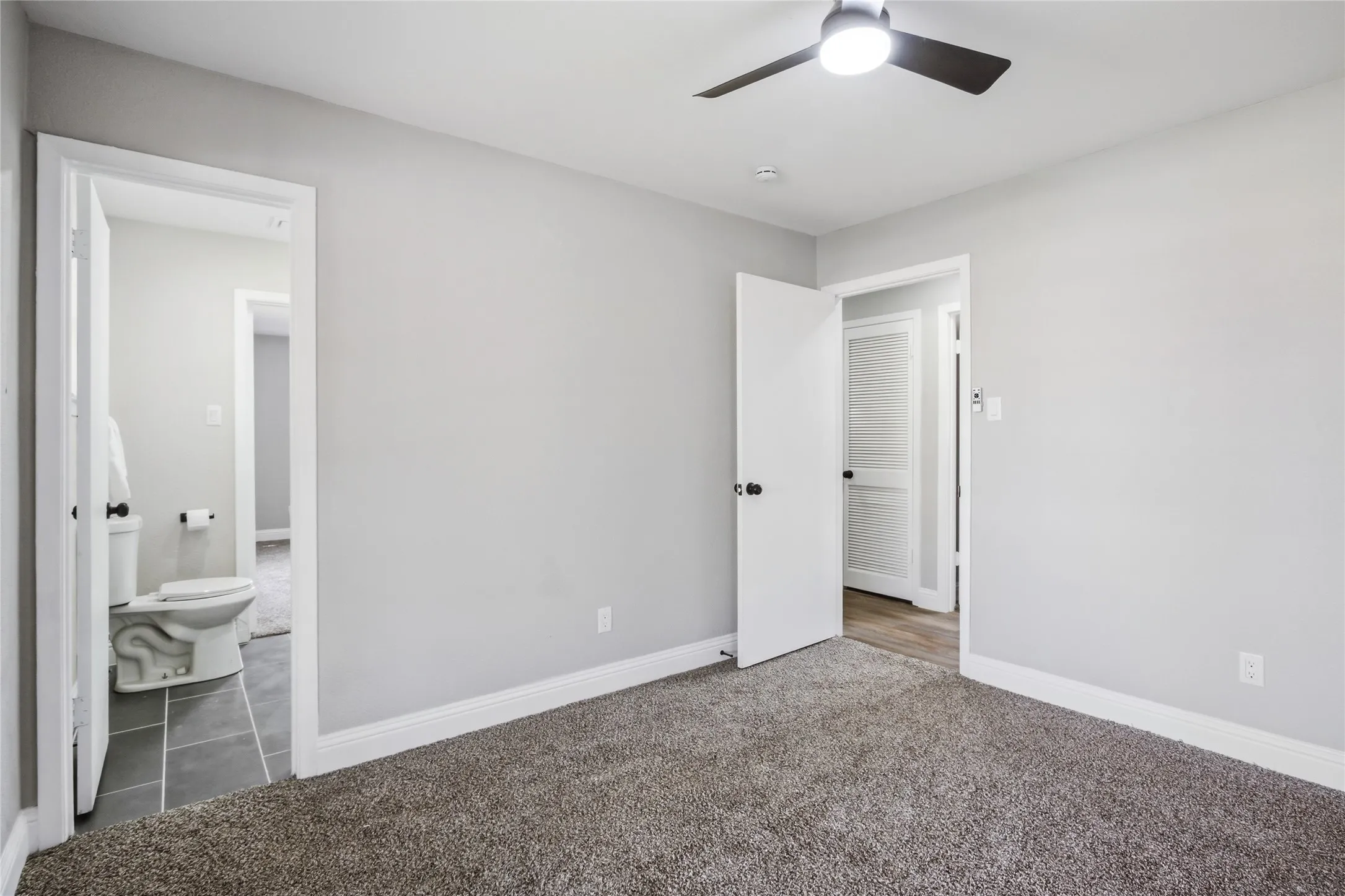 Neutral gray wall paint, dark carpet flooring, ceiling fan with integrated lighting, white trim, and white doors with black hardware