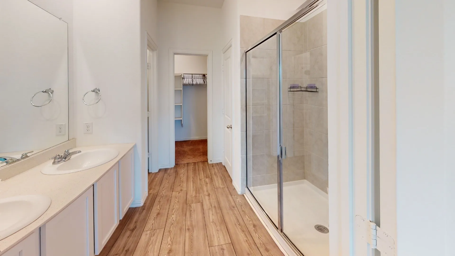 Double vanity with white cabinetry, wood-finish flooring, glass-enclosed shower, and a walk-in closet with built-in shelving