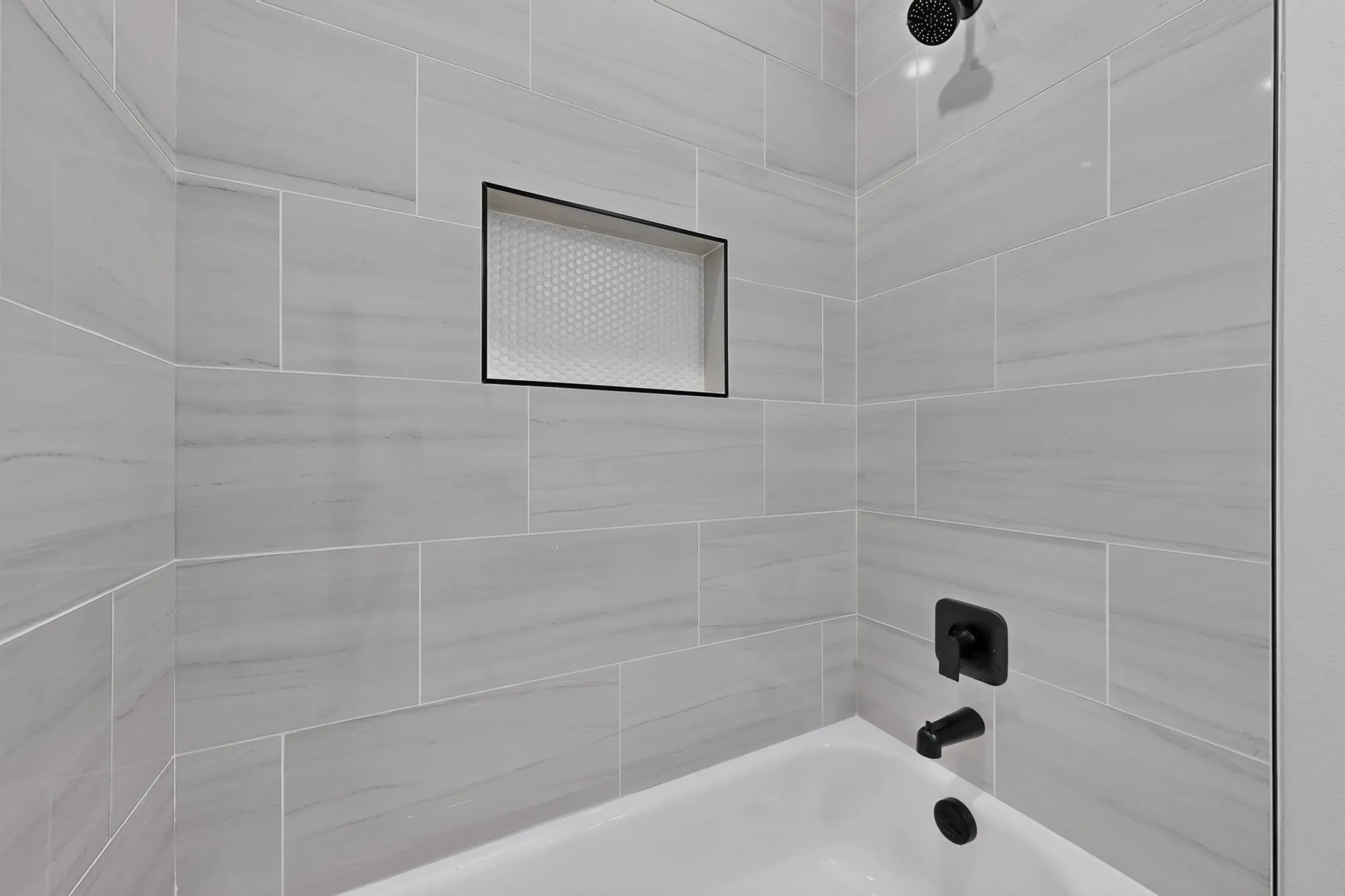 Bathtub with matte black fixtures, an integrated shower niche with honeycomb tile, and light gray subway tile surround