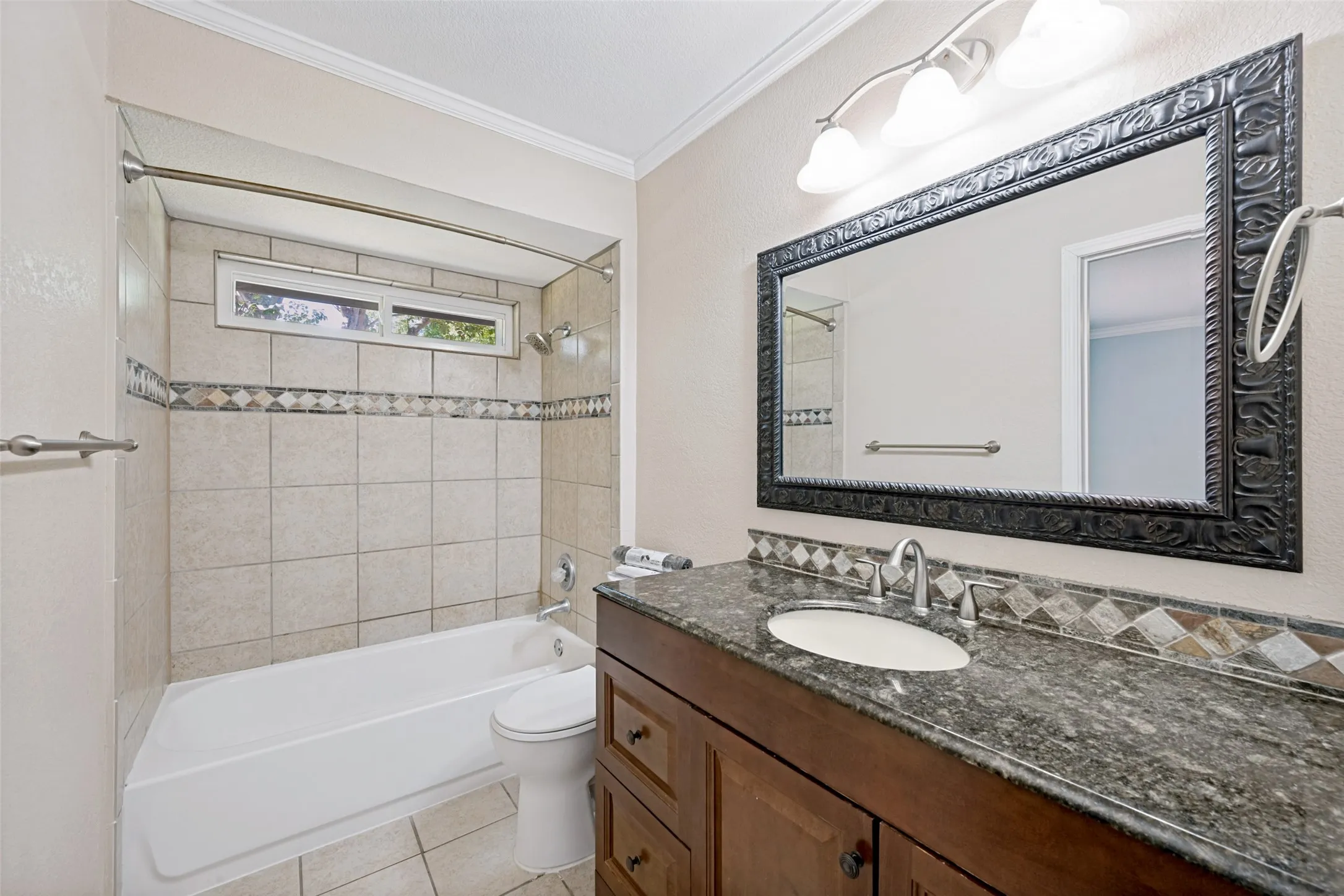Bathroom featuring a wood vanity with a stone countertop, a decorative framed mirror, a tiled shower/tub combination, and a horizontal window
