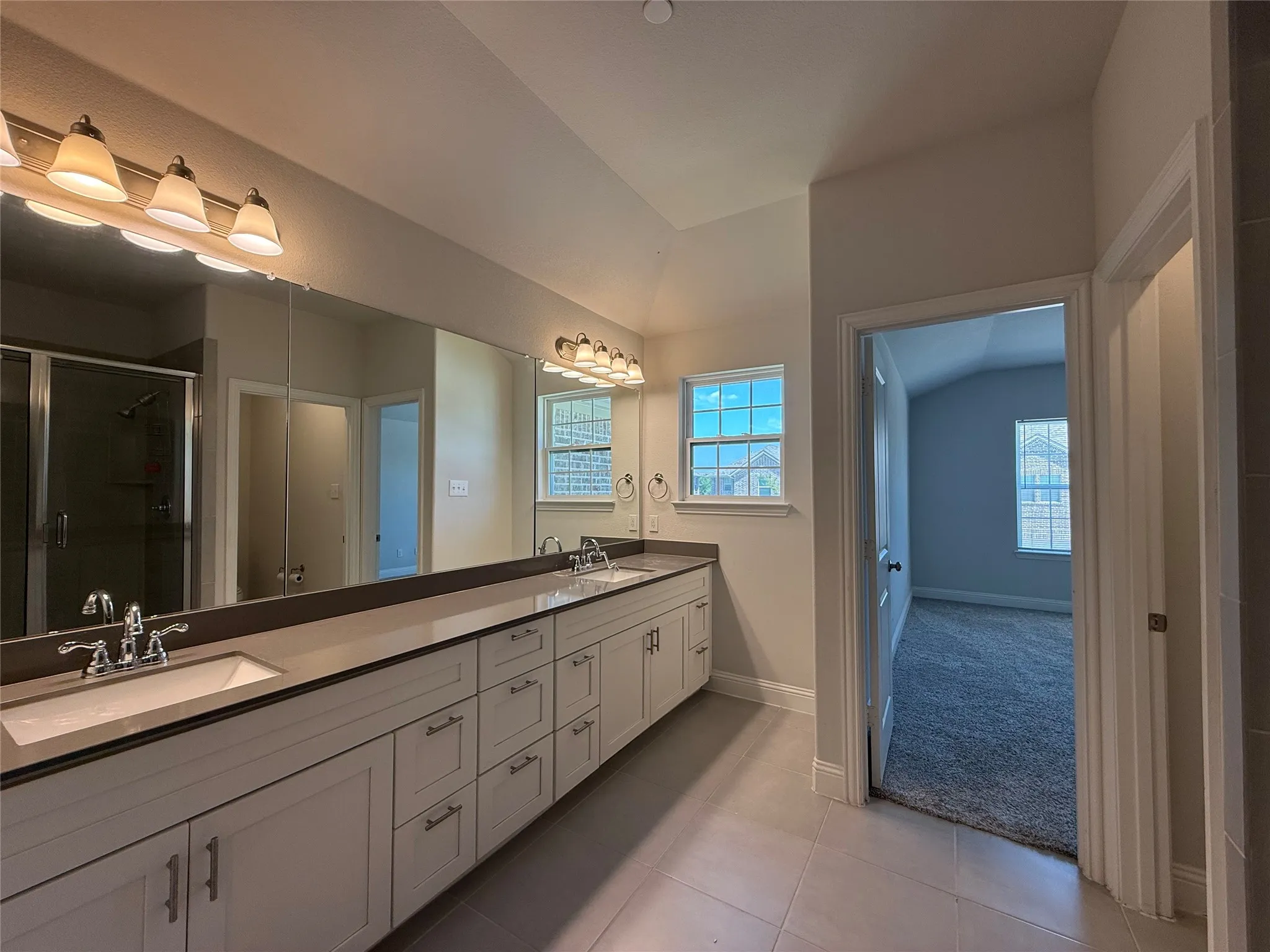 Double vanity with integrated sinks, extensive countertop space, light wood-finish cabinetry, and a walk-in shower with a glass enclosure