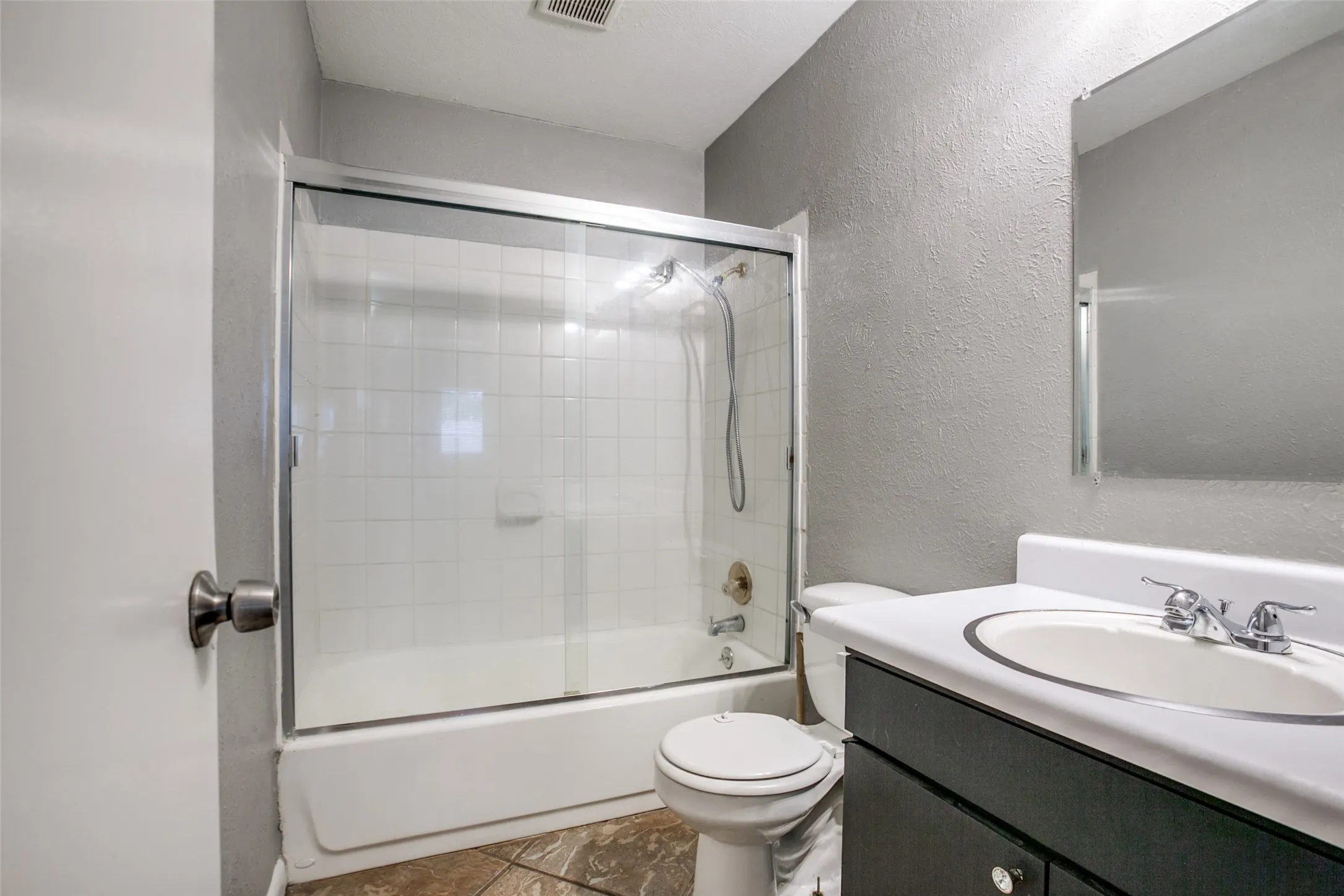Bathroom featuring a shower-tub combination with sliding glass doors, white tiled surround, and a vanity with an integrated sink and dark cabinetry