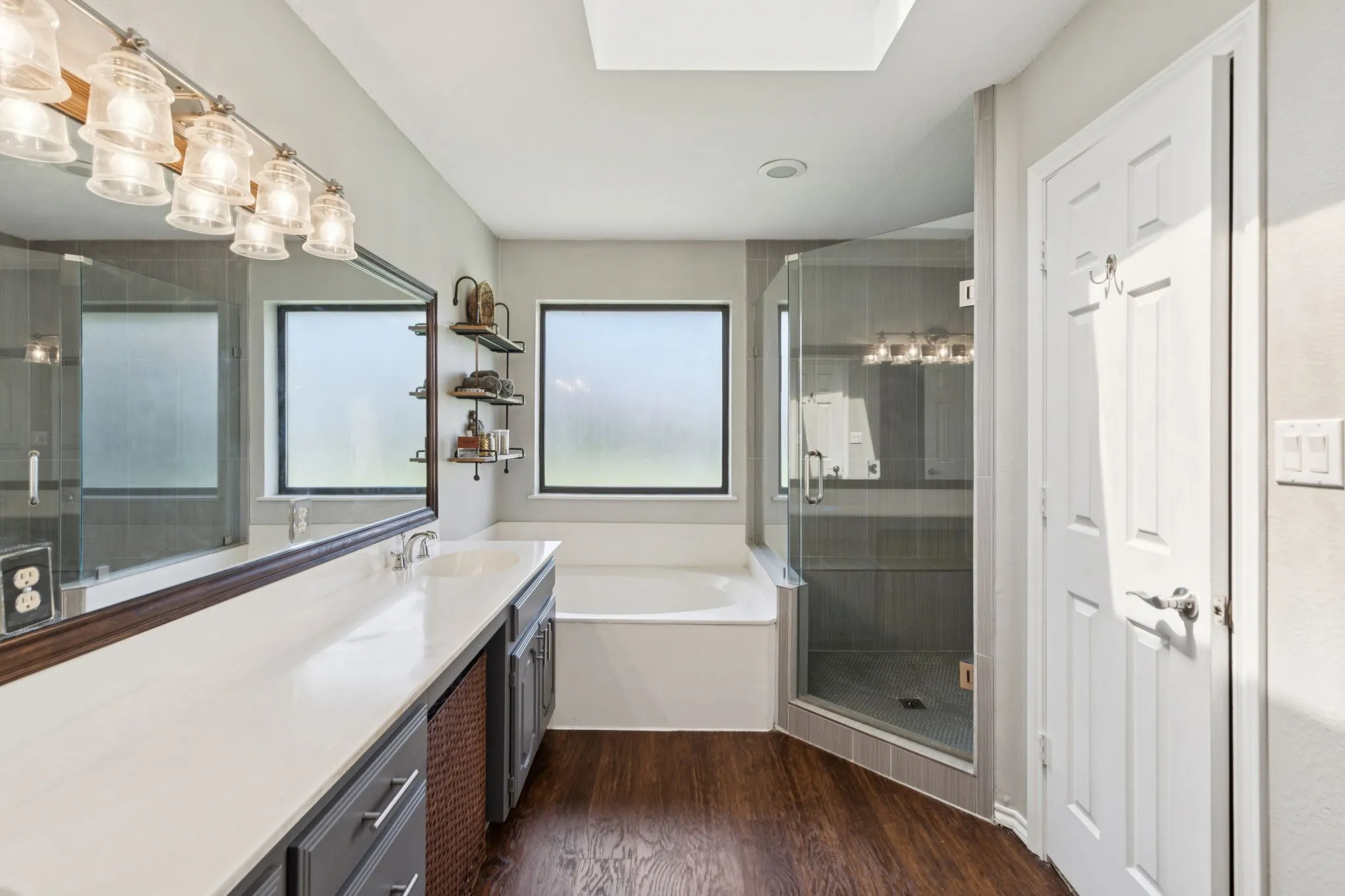 Double vanity with light-tone countertop, glass-enclosed shower, built-in bathtub, and wood-finish flooring