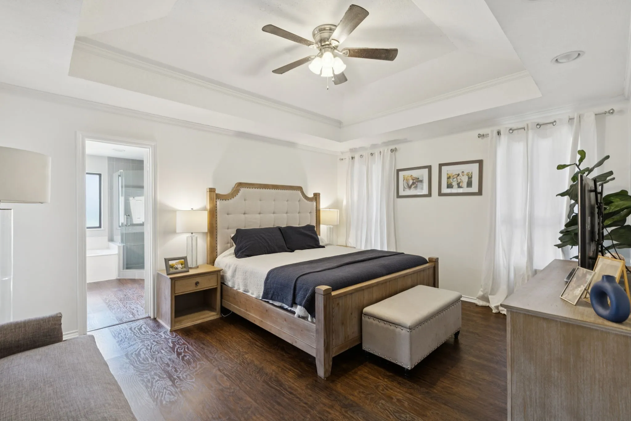 Tray ceiling with crown molding, ceiling fan, wood-finish flooring, recessed lighting, and a doorway opening to a bathroom with a soaking tub and glass-enclosed shower