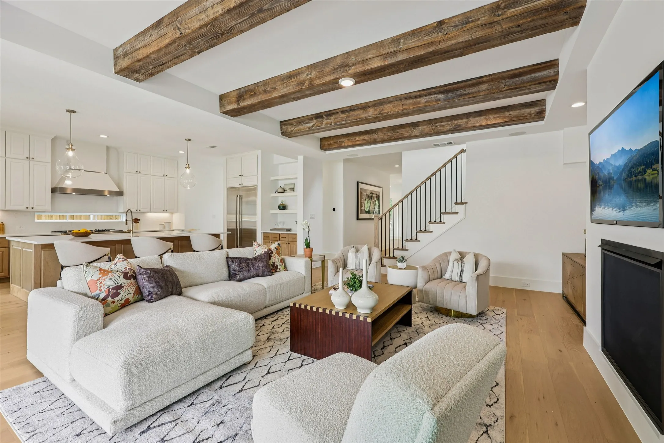 Exposed wood beams, integrated kitchen with island seating, stainless steel appliances, open shelving, and a prominent staircase with metal balusters