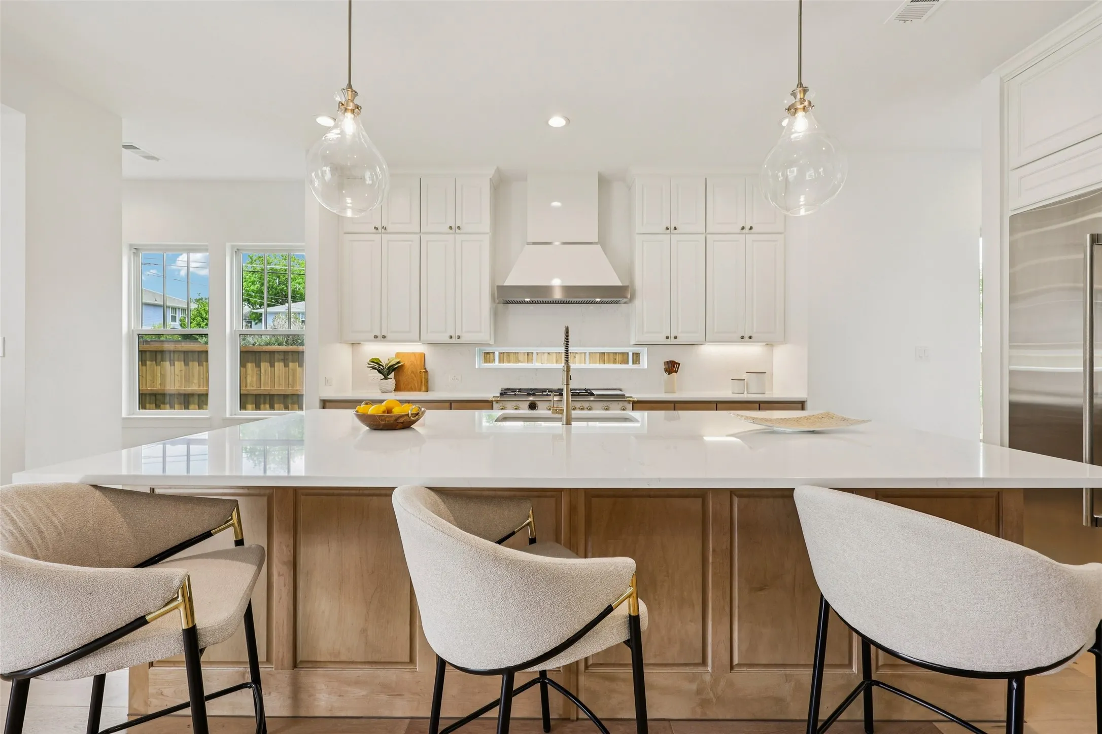 Spacious kitchen island with a light-toned countertop, white upper cabinetry, stainless steel range hood, stainless steel refrigerator, and pendant lighting fixtures