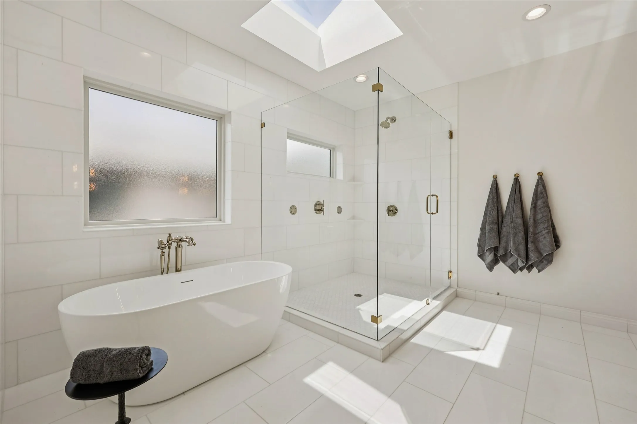 Freestanding soaking tub with floor-mounted faucet, clear glass shower enclosure with brass-finish hardware, subway tile shower surround with recessed niches, large format tile flooring, and a ceiling-mounted skylight