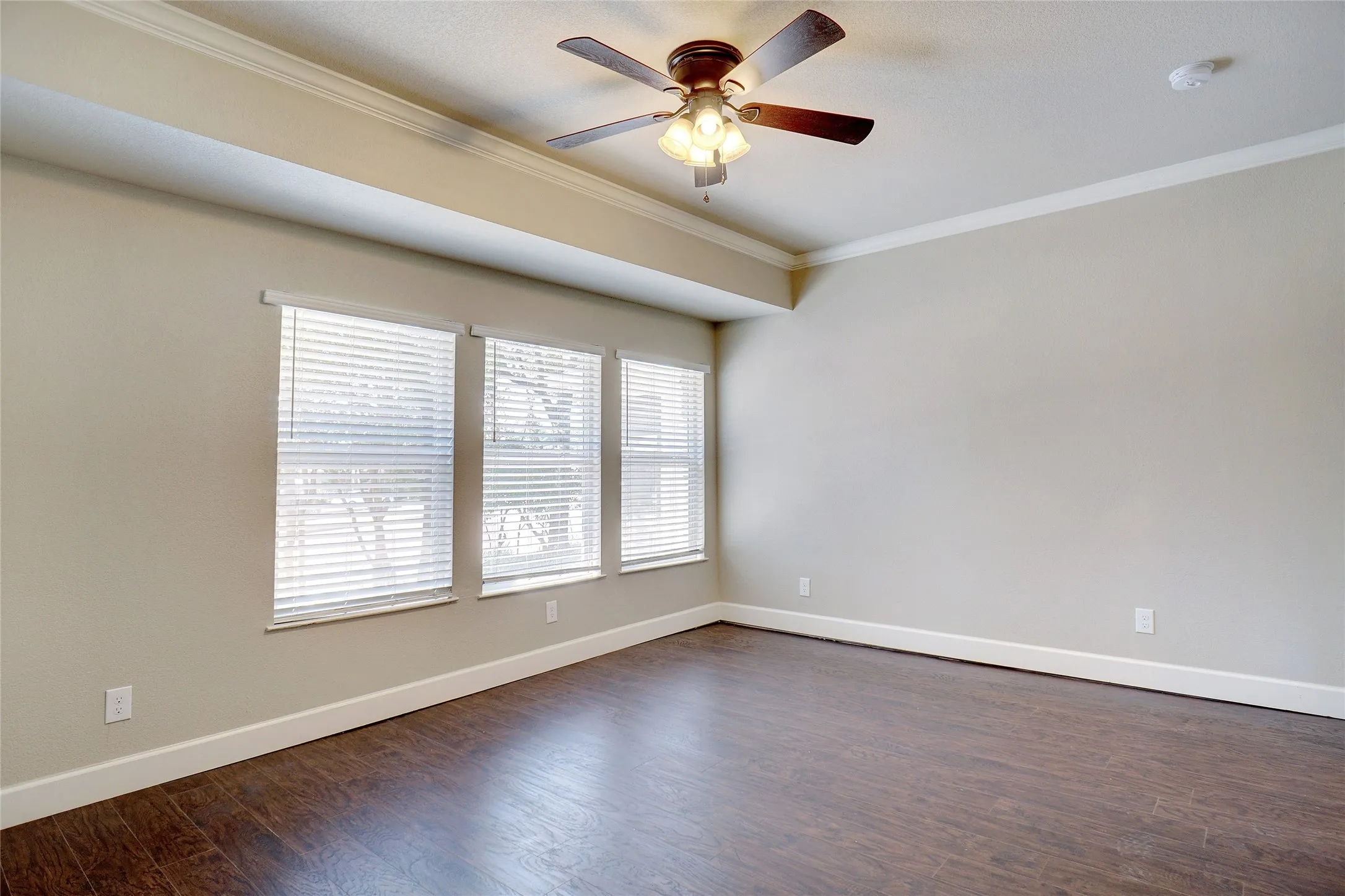 Wood-finish flooring, crown molding, three windows with blinds, and a ceiling fan with integrated lighting