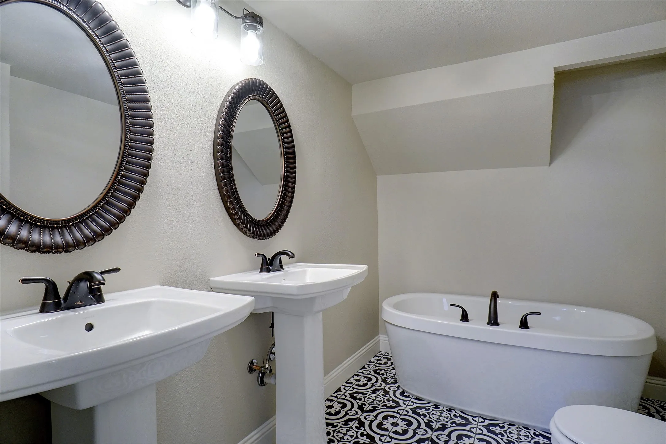 Dual white pedestal sinks with dark bronze fixtures, ornate framed oval mirrors, a freestanding soaking tub, and black and white patterned floor tile