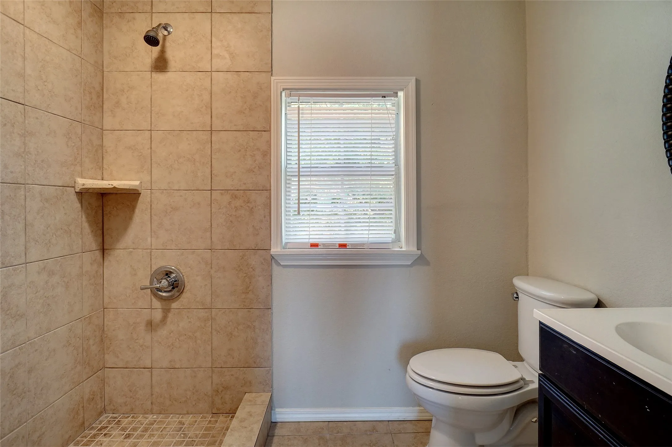 Tiled shower stall with a built-in shelf, chrome fixtures, and a window with blinds
