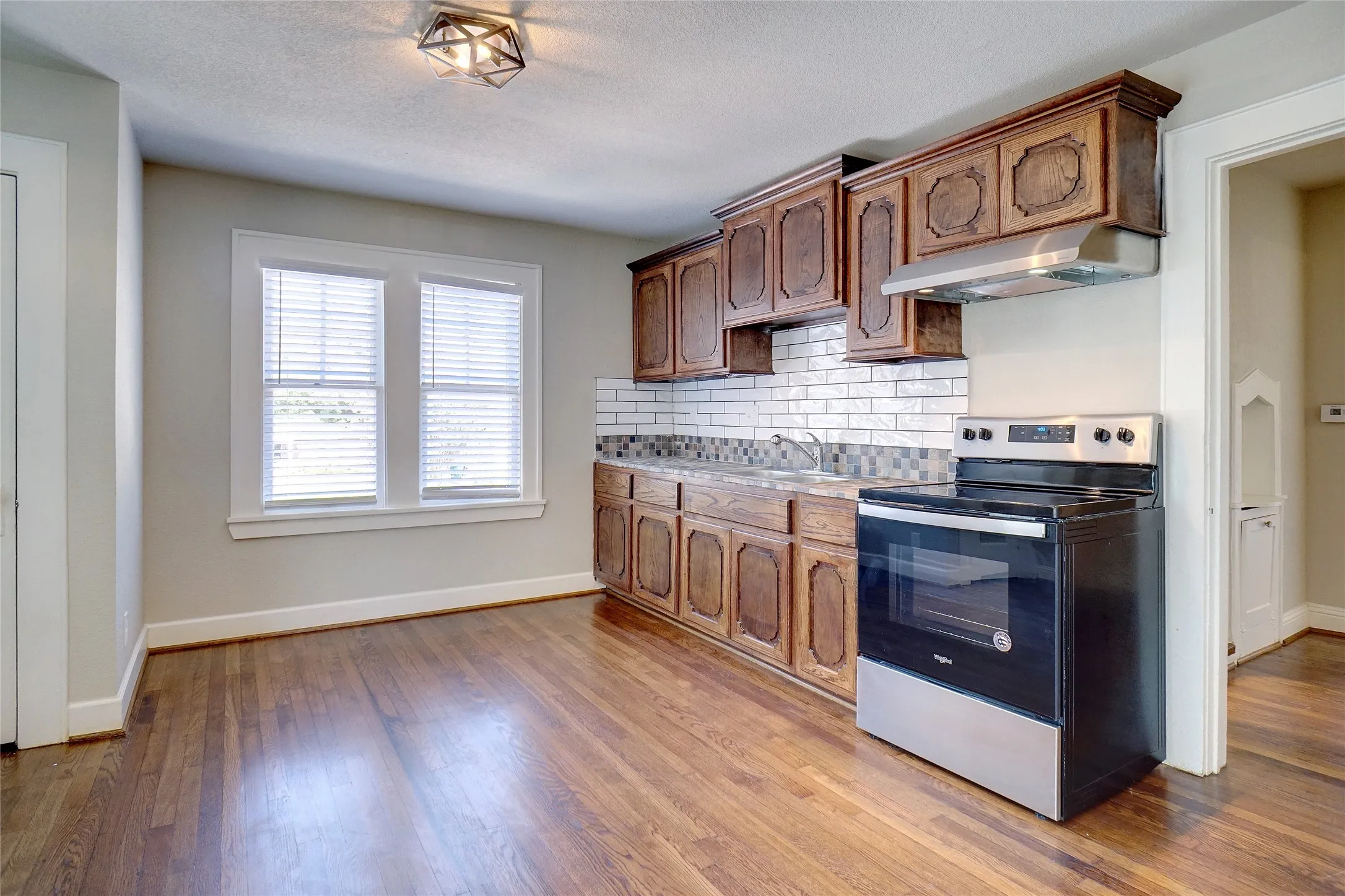Wood-finish flooring, wood cabinetry, subway tile backsplash, stainless steel range with hood, and dual windows with blinds