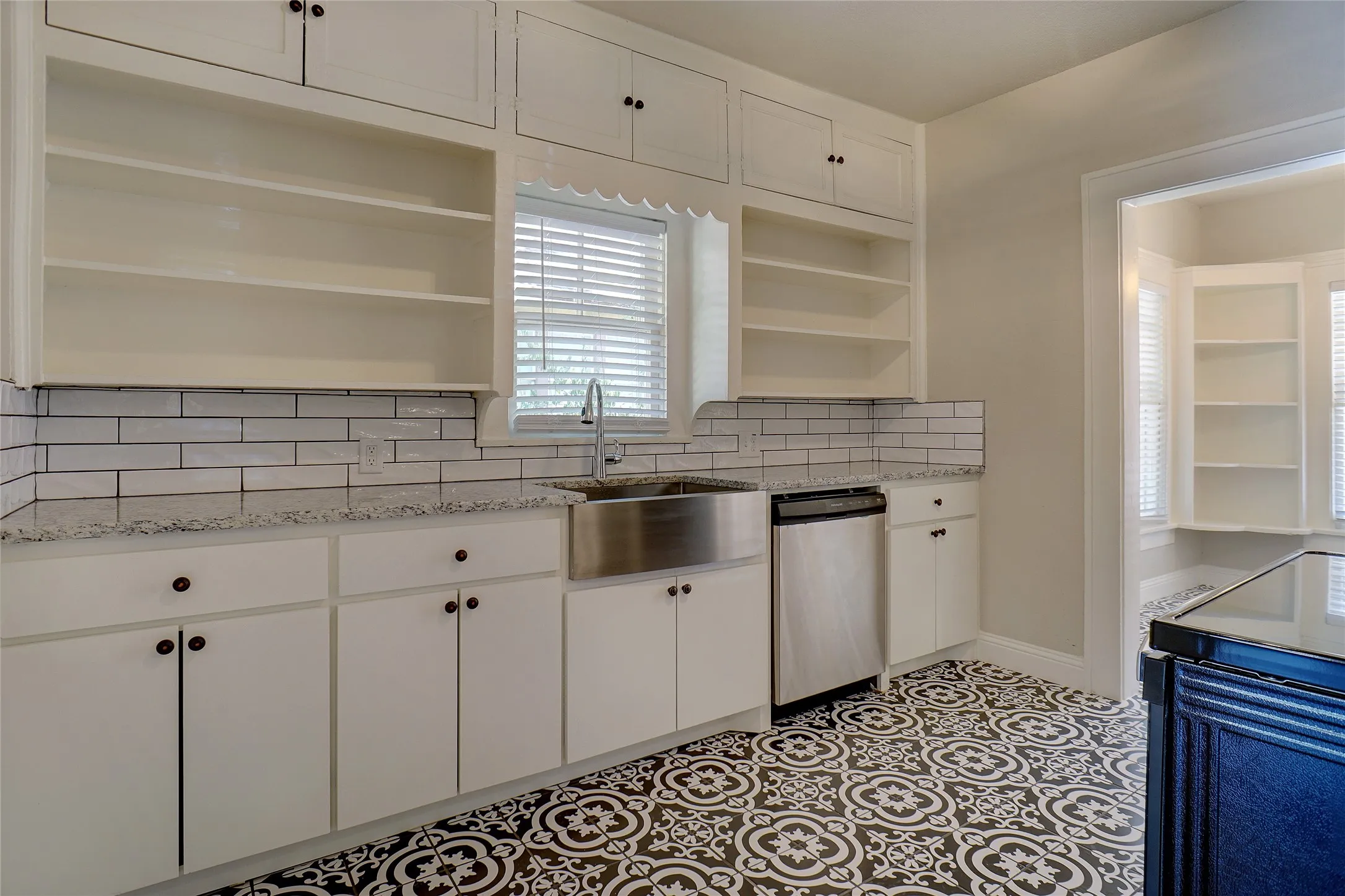 White cabinetry with integrated open shelving, stainless steel farmhouse sink, white subway tile backsplash, light-toned countertops, and patterned floor tile
