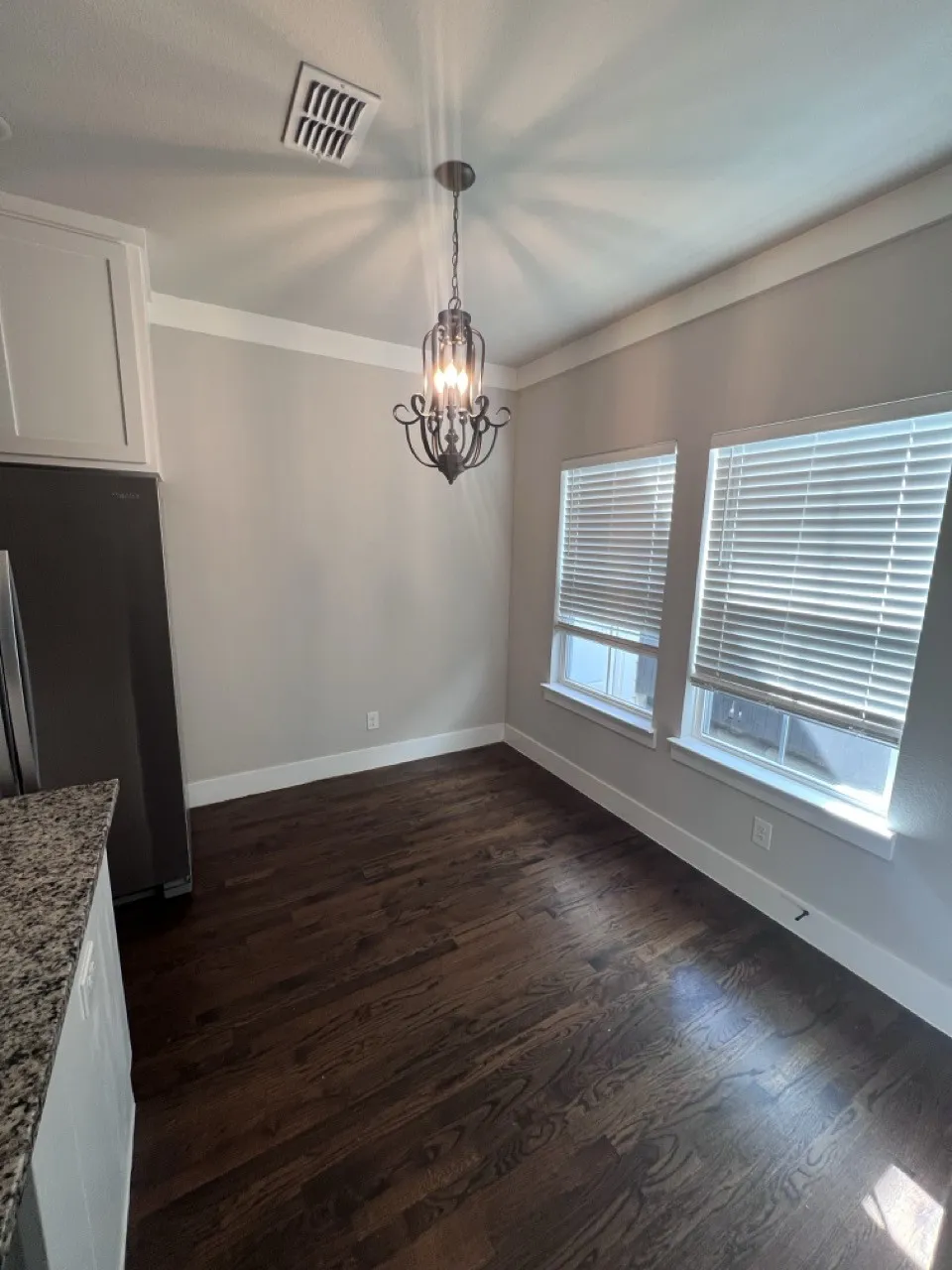 Dark wood-finish flooring, two large windows with blinds, crown molding, a decorative chandelier, and light gray walls