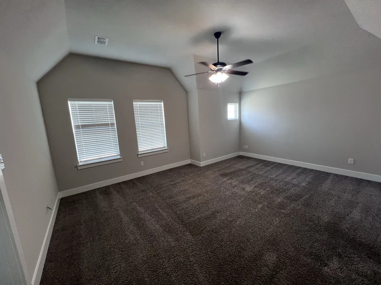 Spacious room featuring vaulted ceilings, dark carpet flooring, multiple windows with blinds, and a ceiling fan with integrated lighting