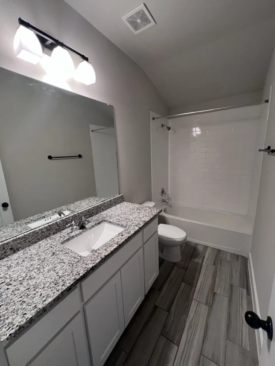 Granite-style countertop with integrated sink, white shaker-style vanity cabinetry, wood-finish tile flooring, recessed bathtub with tile surround, and contemporary vanity lighting