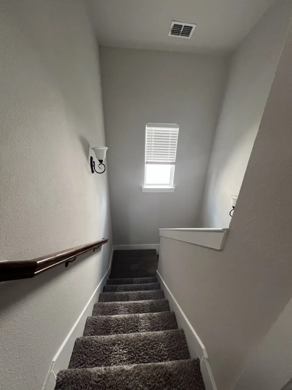 Carpeted staircase with a dark wood-finish handrail, wall-mounted sconce lighting, and a small window featuring horizontal blinds