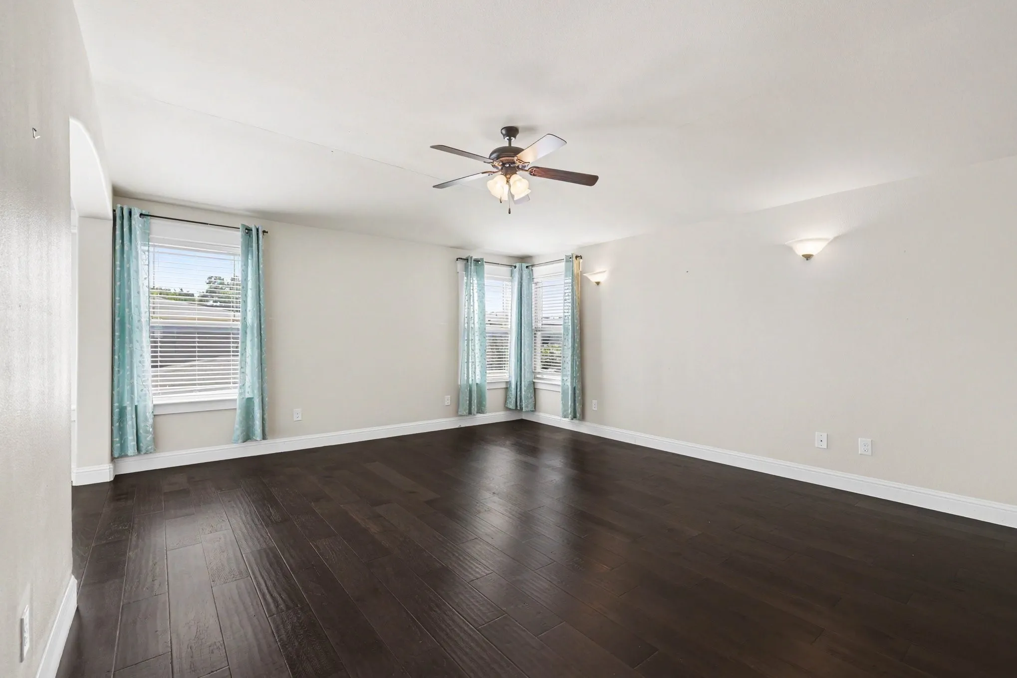 Dark wood-finish flooring, multiple windows with blinds, ceiling fan with integrated lighting, and wall sconces