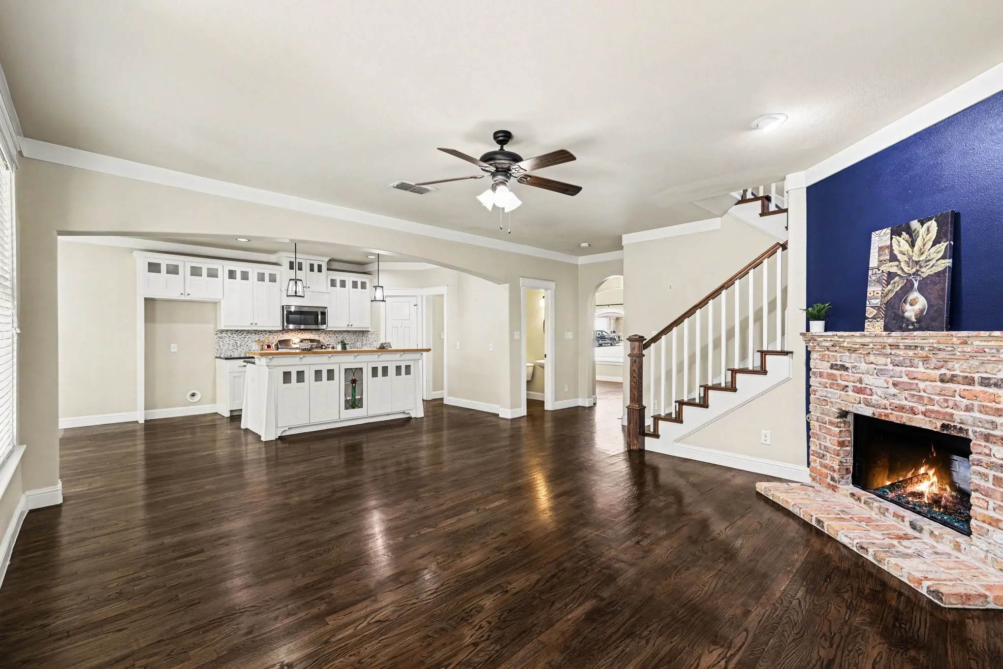 Wood-finish flooring throughout, a brick fireplace, and a kitchen featuring white cabinetry, a central island, and pendant lighting