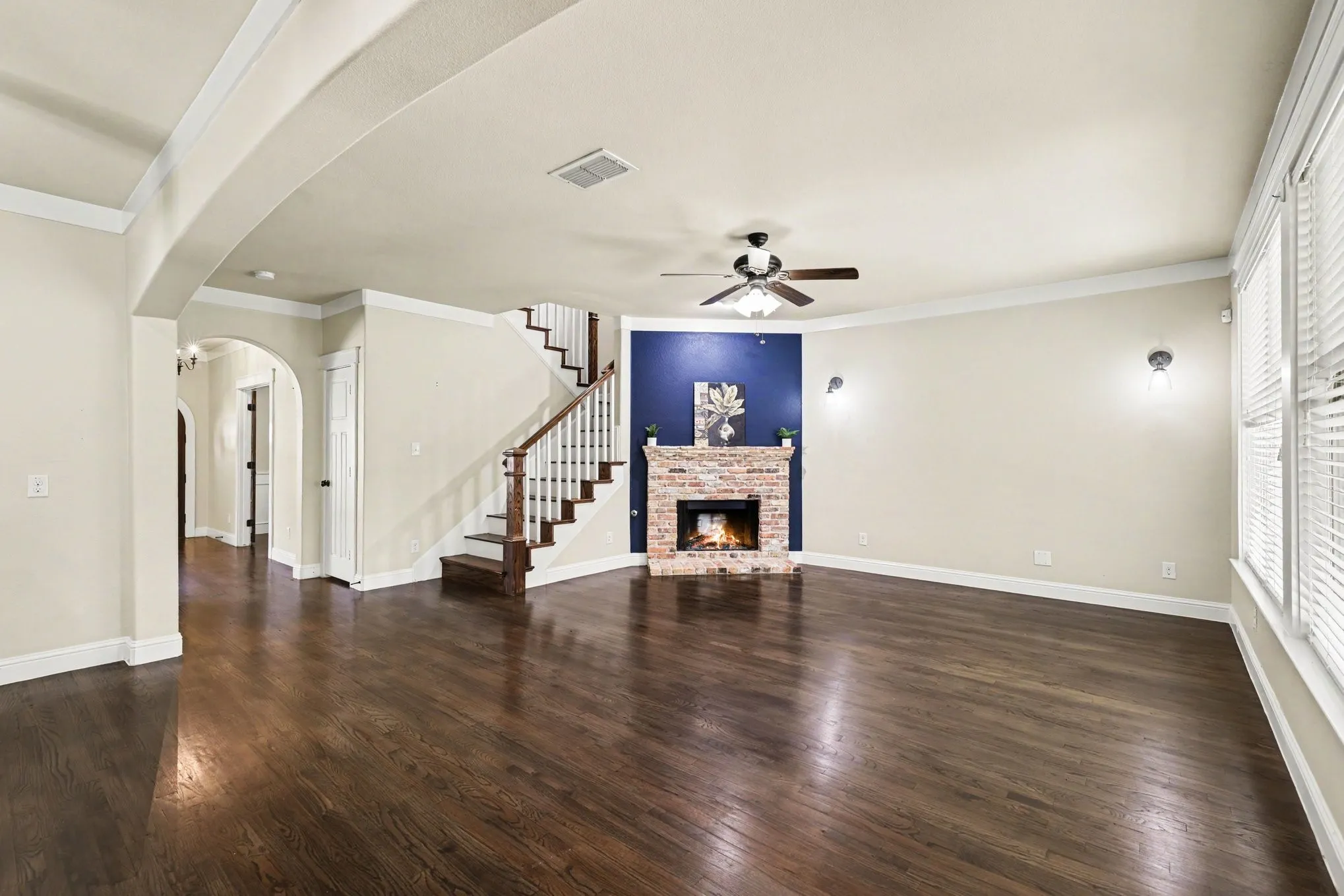 Rich wood-finish flooring throughout, brick-surround fireplace with hearth, white window blinds, and a staircase with wood treads and white risers