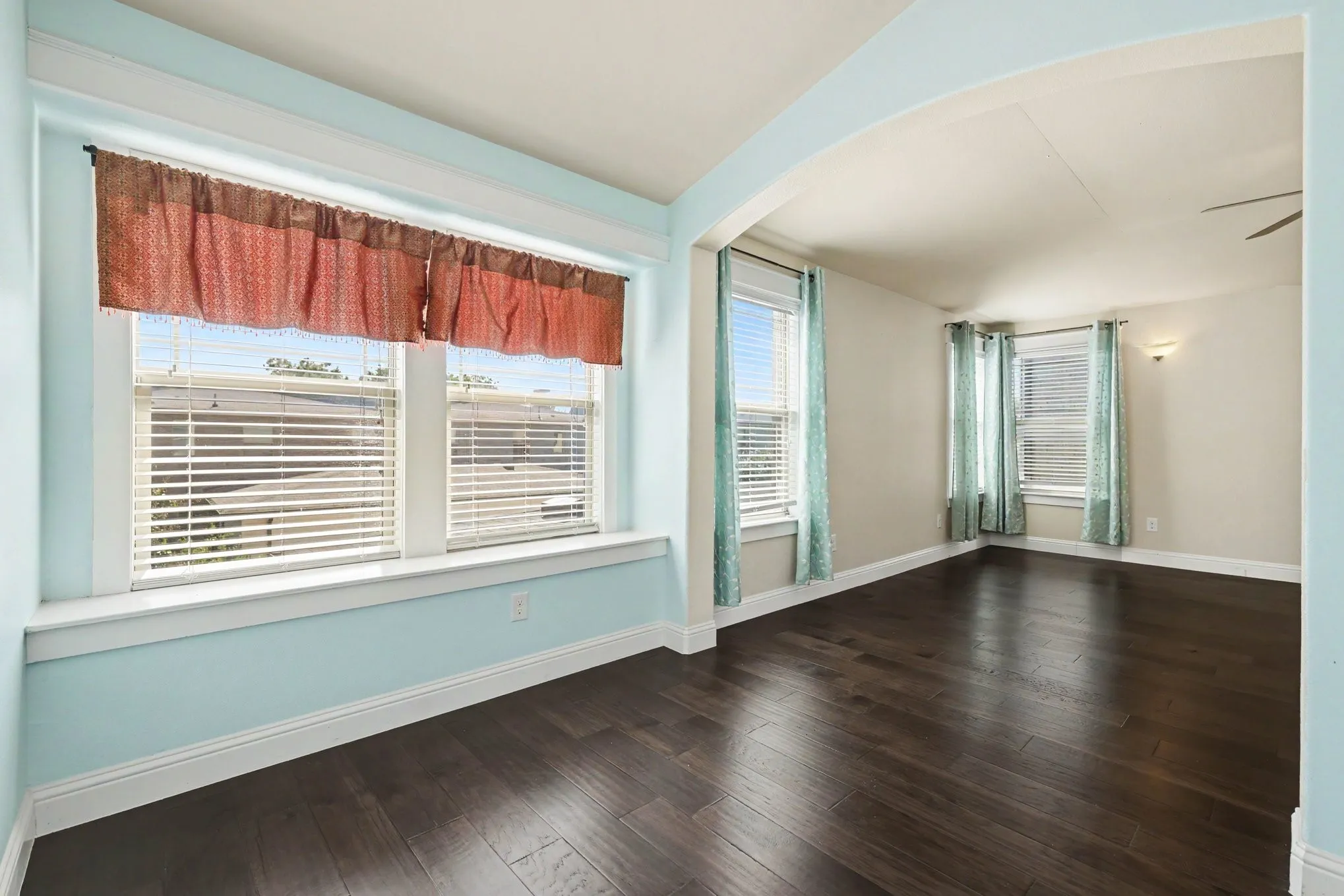 Dark wood-finish flooring throughout, featuring a wide arched doorway, multiple windows with blinds, a built-in window ledge, and a ceiling fan