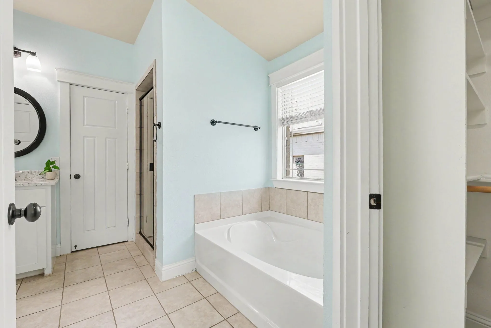 Tiled flooring, built-in bathtub with tiled surround, freestanding vanity with integrated sink, circular wall-mounted mirror, and a glass-enclosed shower