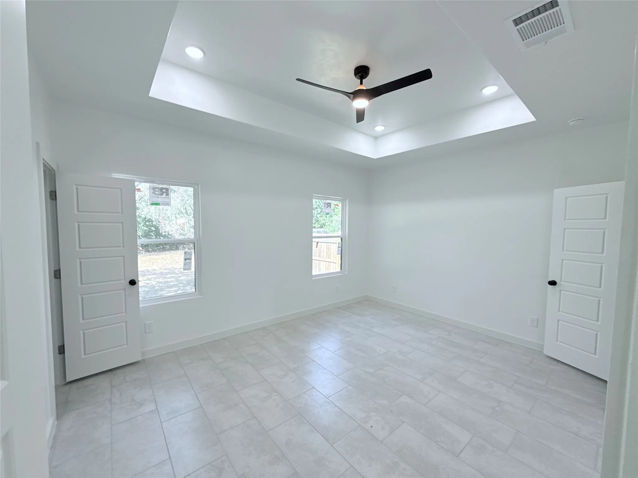 Recessed tray ceiling with integrated lighting, contemporary ceiling fan, dual windows, paneled doors with matte black hardware, and large format tile flooring