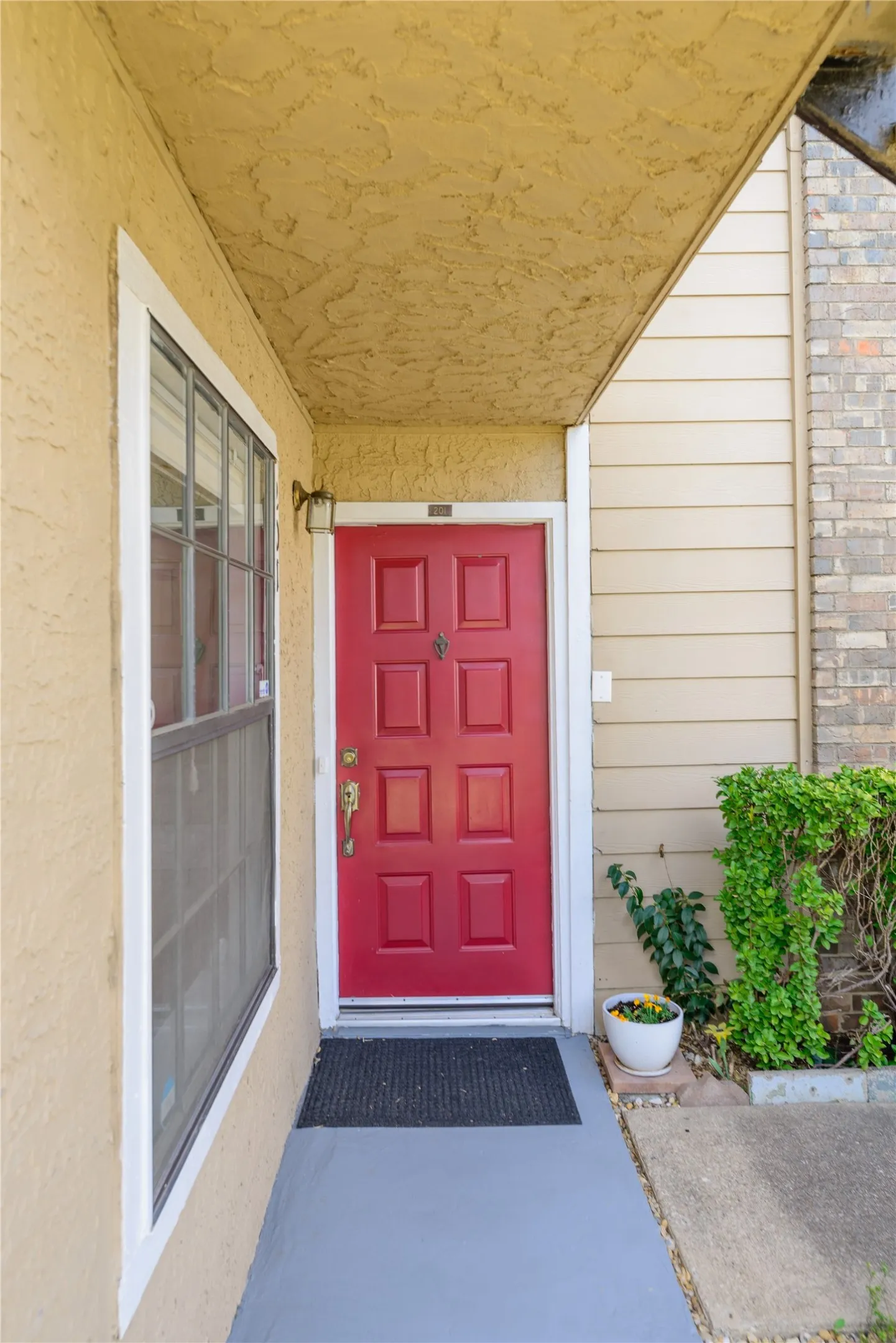 Red paneled entry door with brass hardware, stucco exterior, covered entryway, and an exterior wall light fixture