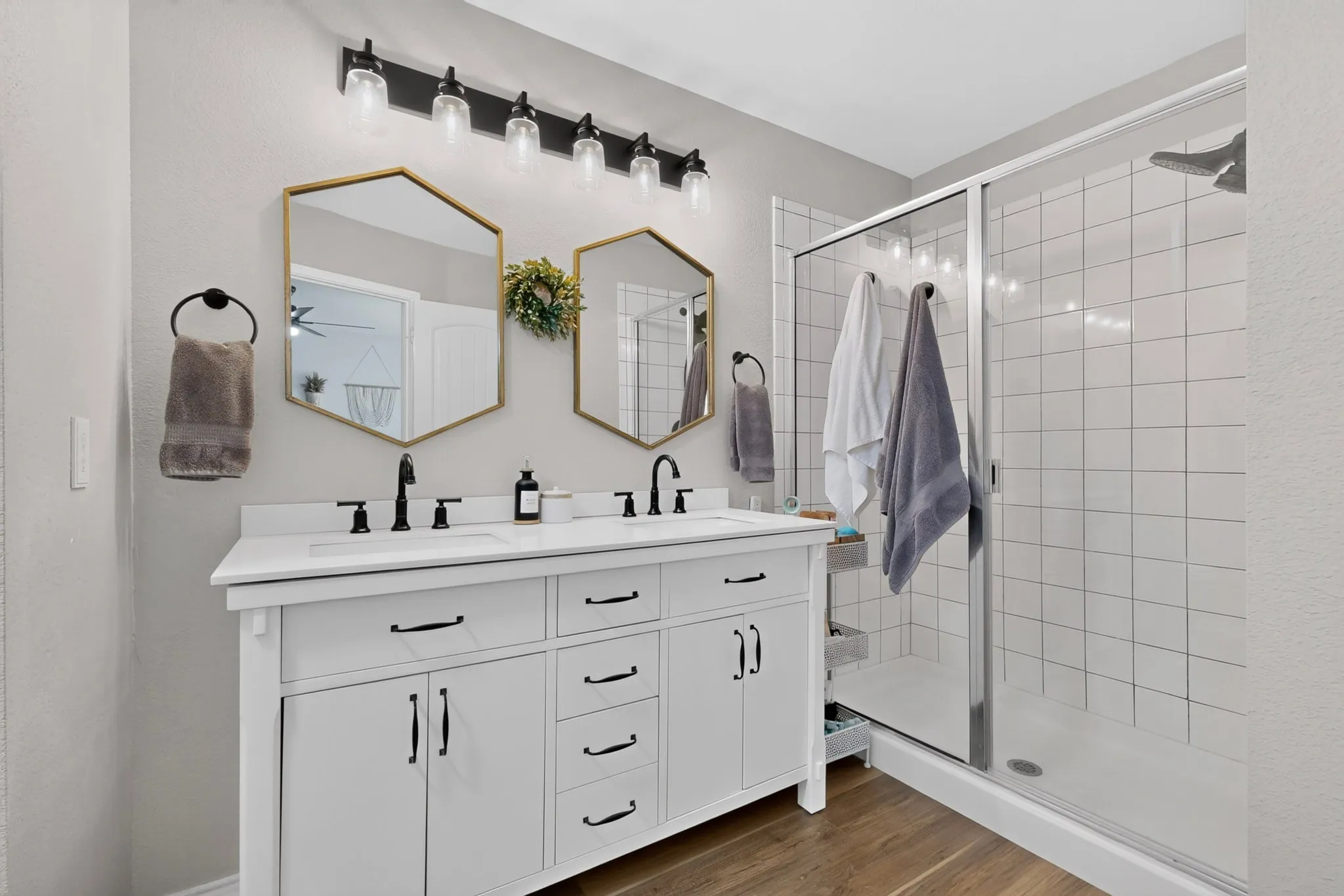 Dual vanity with solid surface countertop, hexagonal mirrors, black hardware, and a glass-enclosed shower with white subway tile