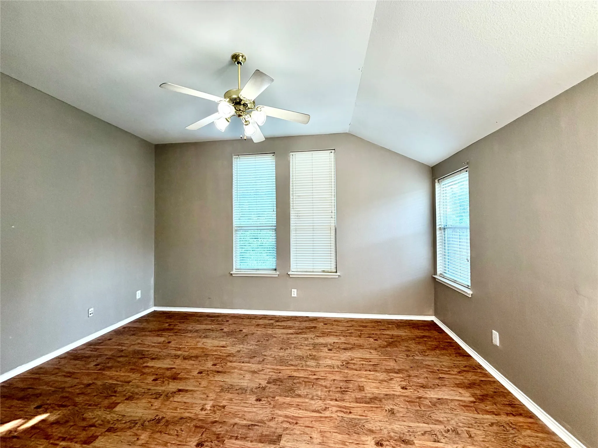 Vaulted ceiling with a brass-finish ceiling fan, three windows with blinds, and wood-finish flooring