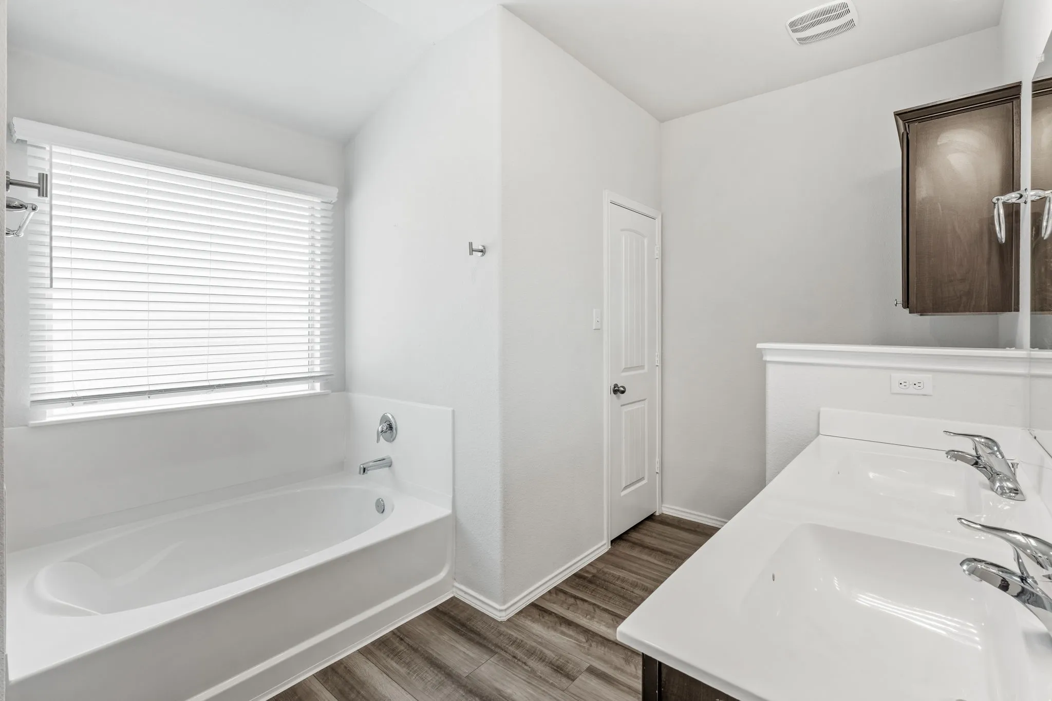 White bathtub with chrome fixtures, wood-finish flooring, dual vanity with integrated sinks, and dark wood-finish cabinetry