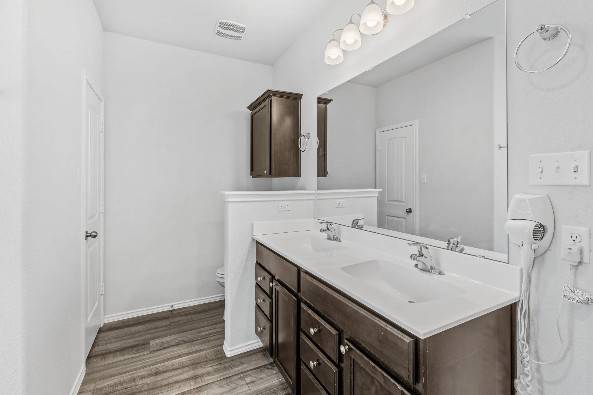 Dual vanity with integrated sinks, wood-finish flooring, dark wood cabinetry, and a large wall-mounted mirror