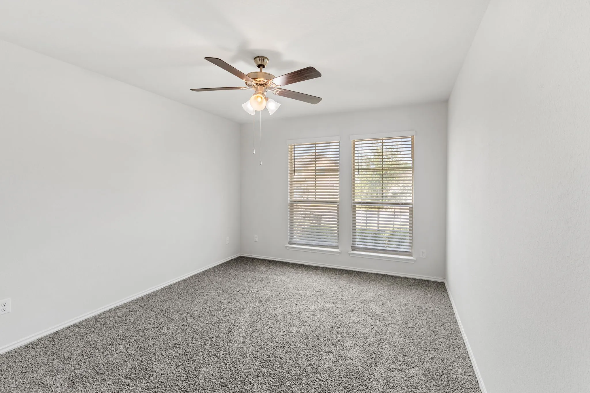 White walls, gray carpeting, two windows with blinds, and a ceiling fan with integrated lighting