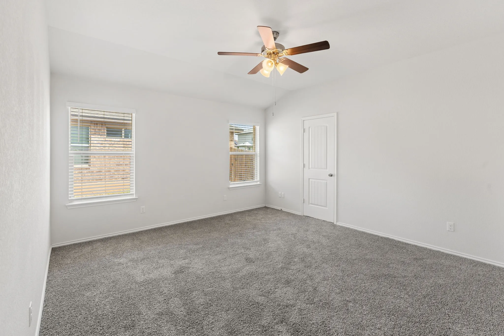 Spacious room featuring neutral carpeting, two windows with blinds, a ceiling fan with integrated lighting, and a paneled door