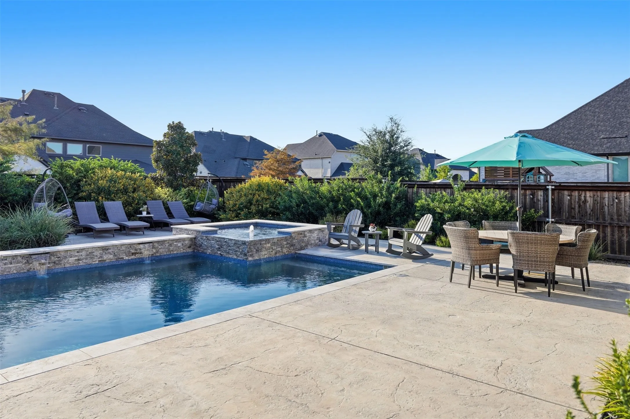 Expansive patio area featuring a built-in swimming pool, a raised spa with a water feature, a stone-clad retaining wall, and mature landscaping