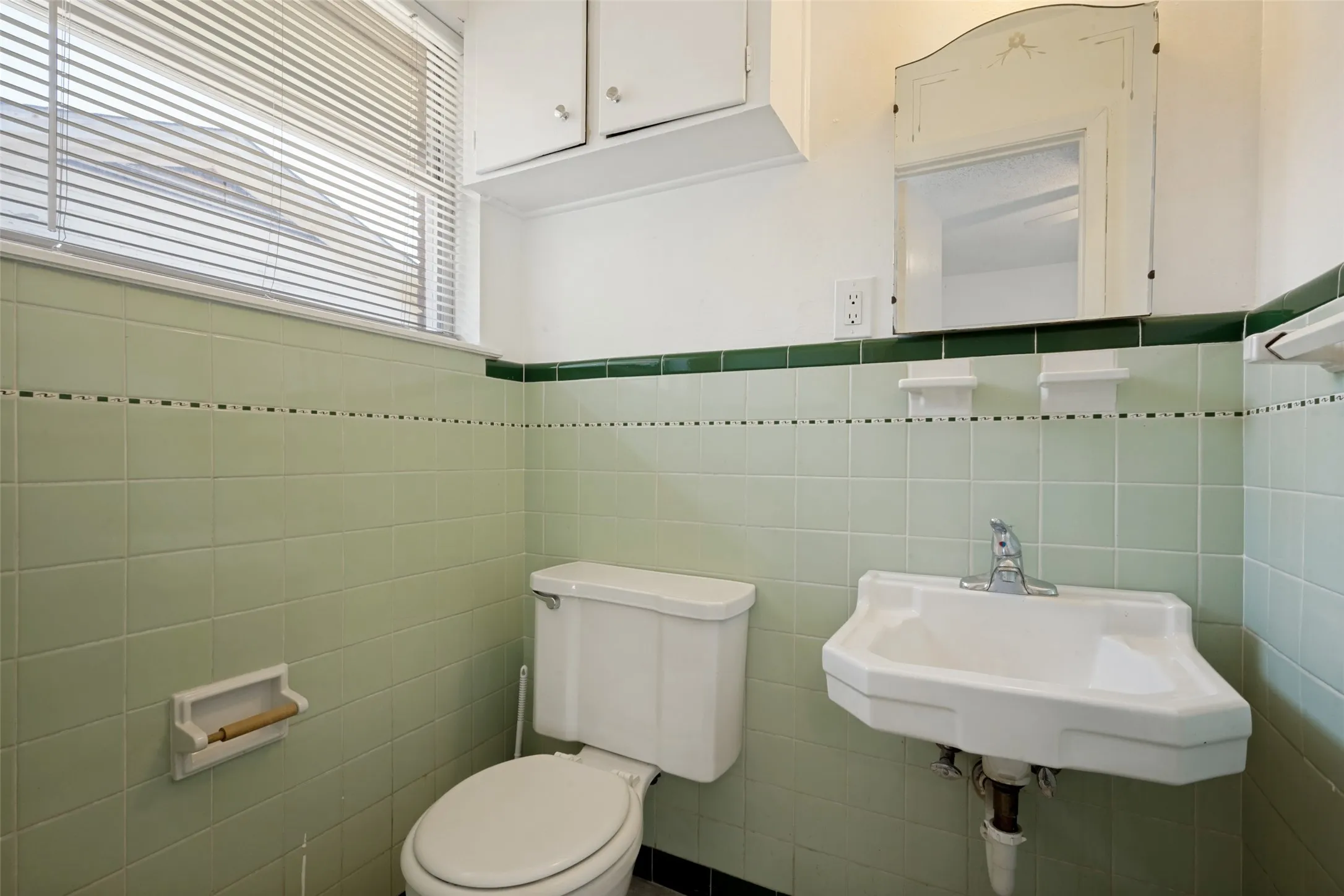 Window with horizontal blinds, light green wall tile with dark green trim, wall-mounted sink with chrome faucet, built-in wall mirror, white toilet