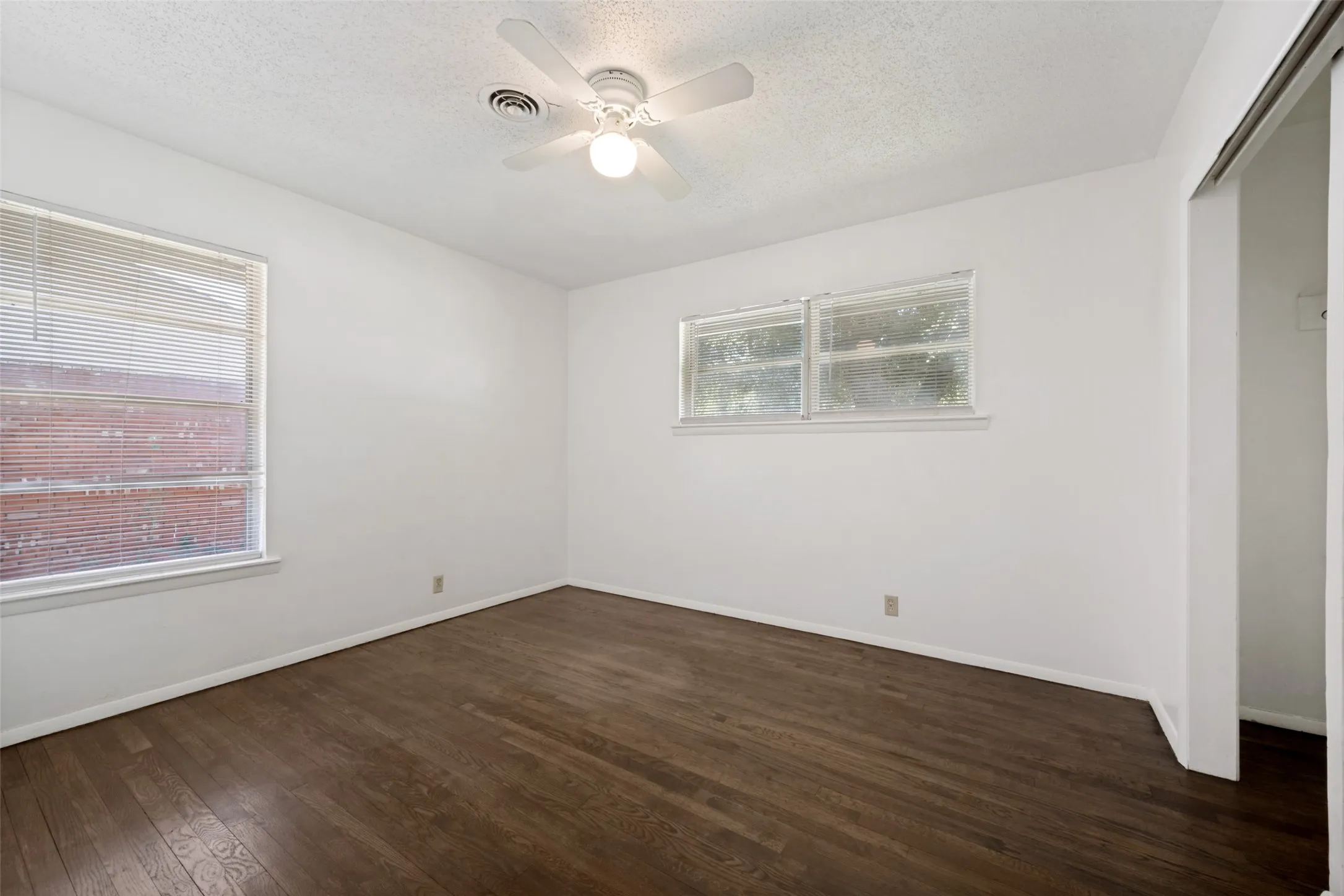 Wood-finish flooring, white walls, two windows with blinds, and a ceiling fan with integrated lighting