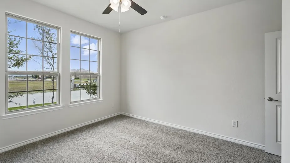 Dual windows with grid patterns, ceiling fan with light fixture, light gray carpeting, neutral wall paint, and a solid interior door with a silver-tone handle