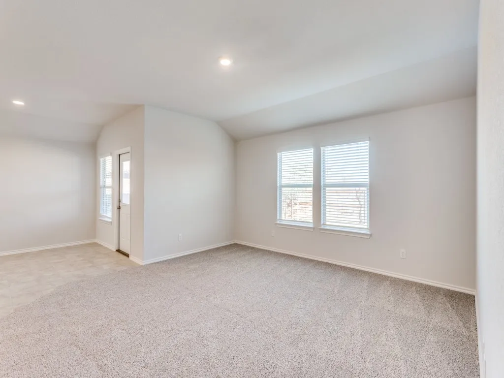 Neutral tone wall paint, recessed ceiling lighting, light-colored carpeting, multiple windows with blinds, and an exterior door with louvered blinds